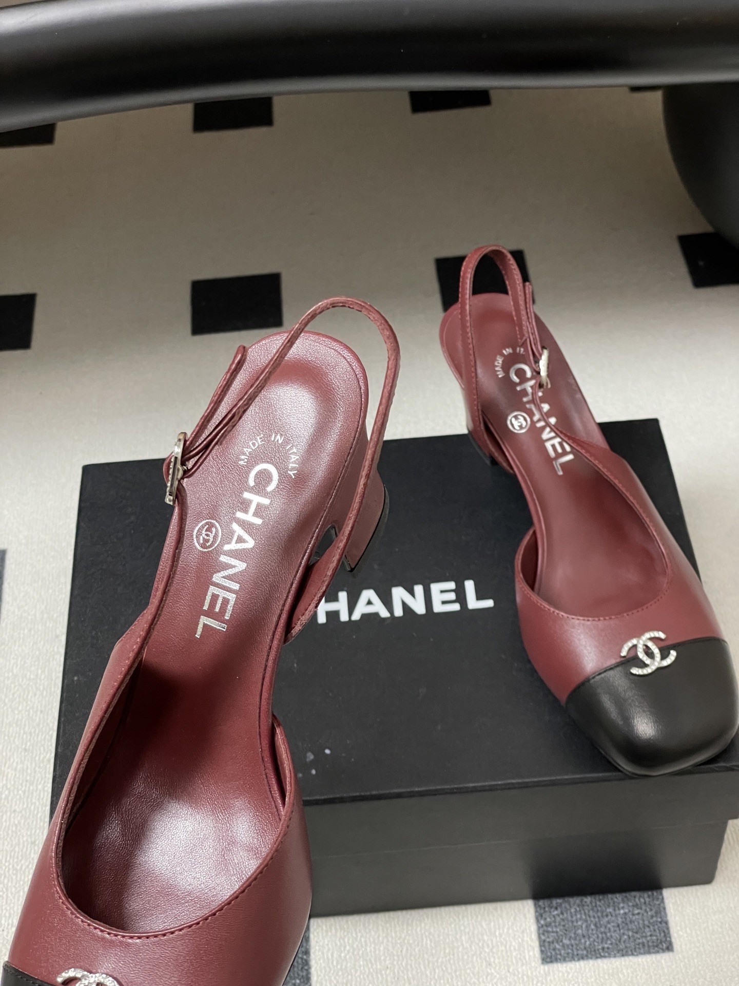 A pair of Chanel burgundy leather slingback heels with black cap toes and crystal-embellished CC logos on a black surface. - Hình 7