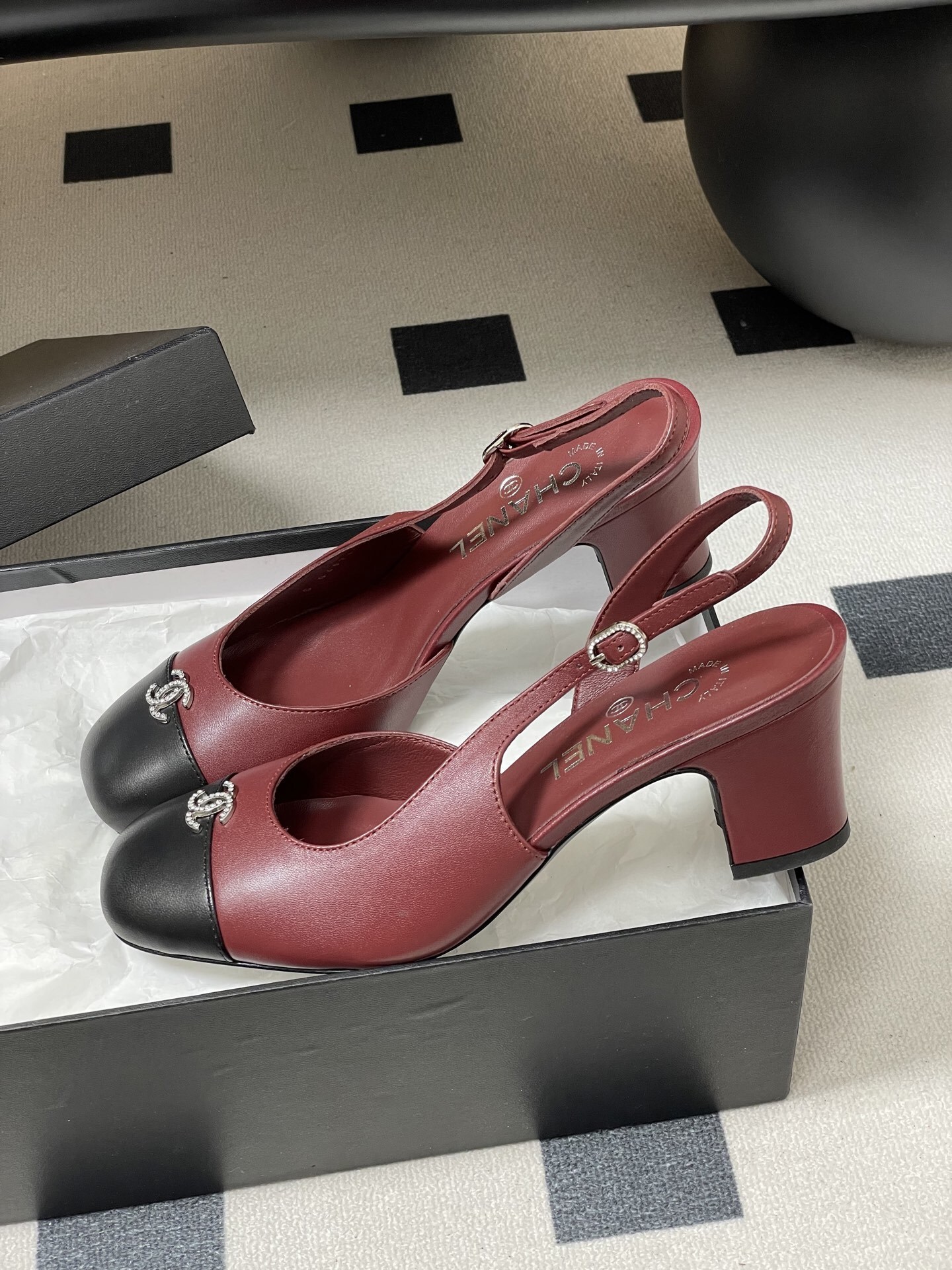 A pair of Chanel burgundy leather slingback heels with black cap toes and crystal-embellished CC logos on a black surface. - Hình 4