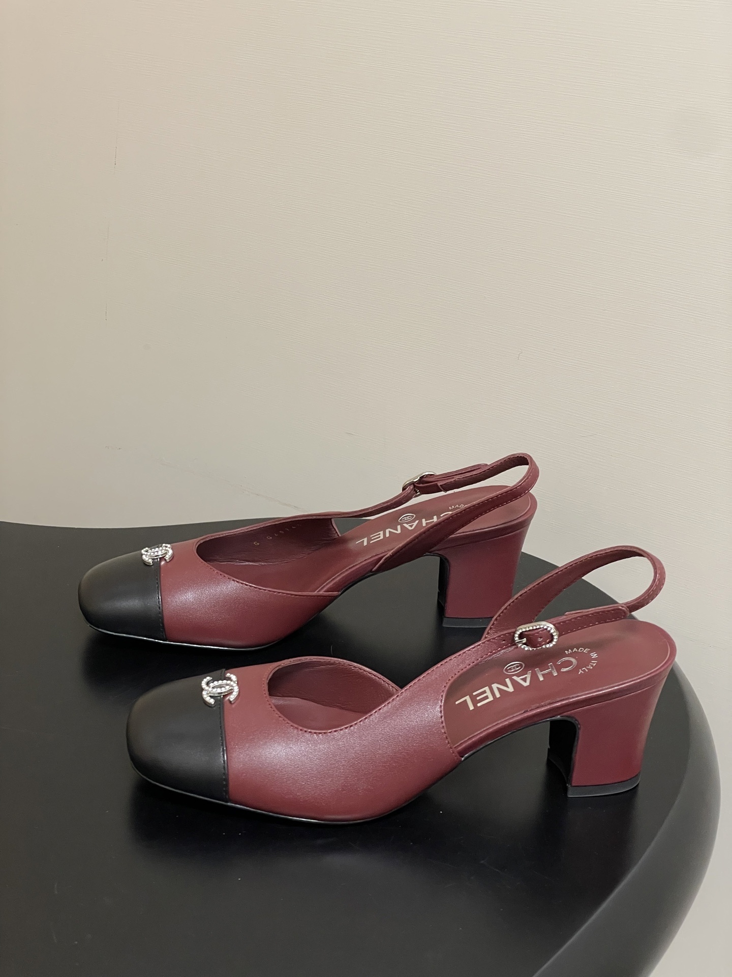 A pair of Chanel burgundy leather slingback heels with black cap toes and crystal-embellished CC logos on a black surface. - Hình 3