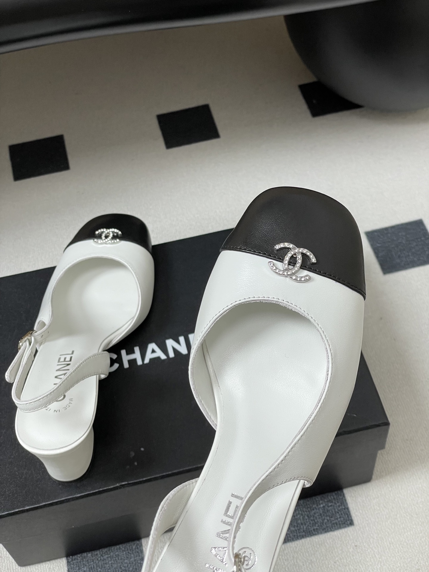 Pair of Chanel white leather slingback pumps with black toe caps and crystal CC logos on a black table. - Hình 9