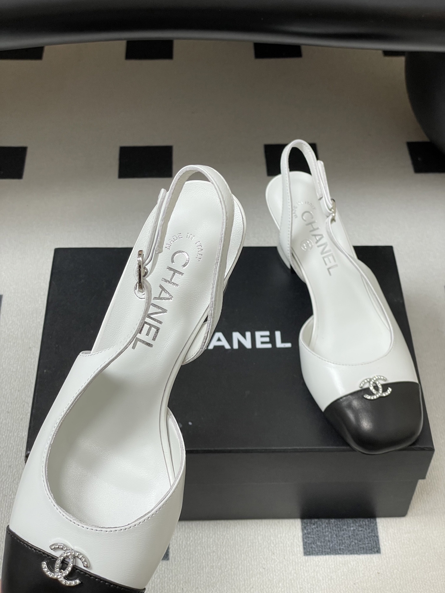 Pair of Chanel white leather slingback pumps with black toe caps and crystal CC logos on a black table. - Hình 7