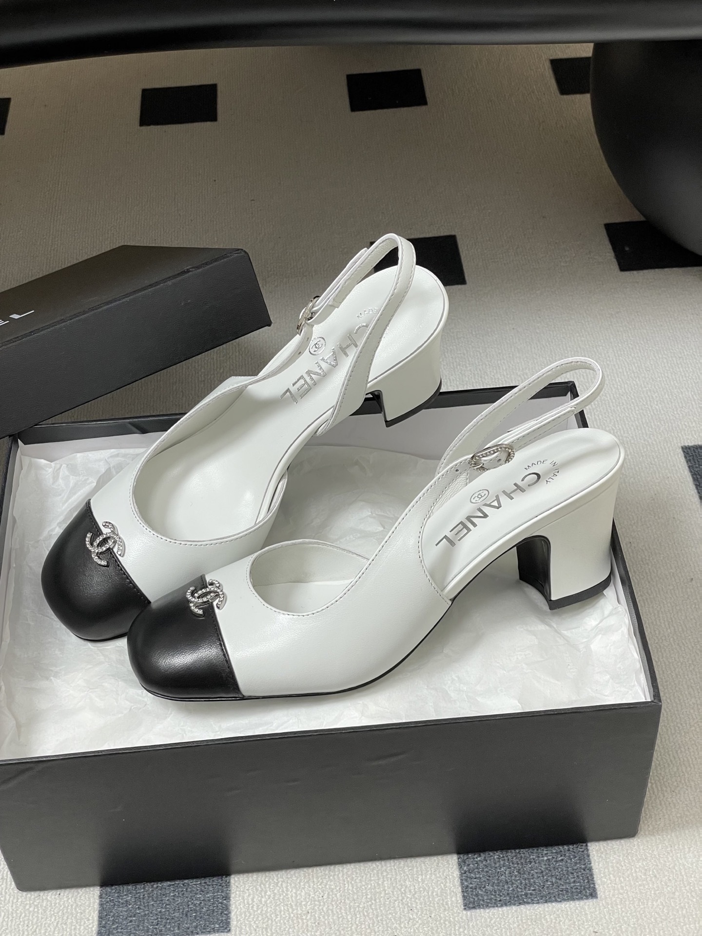 Pair of Chanel white leather slingback pumps with black toe caps and crystal CC logos on a black table. - Hình 4