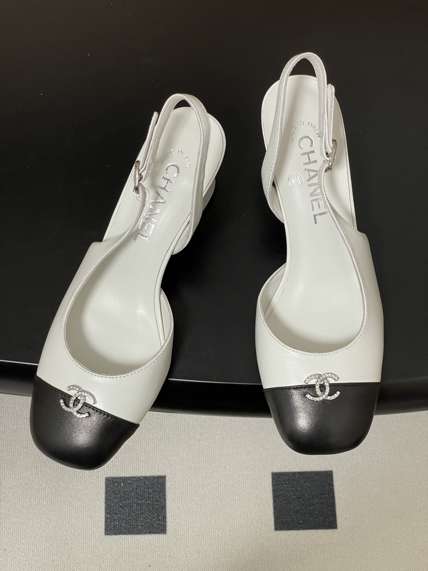 Pair of Chanel white leather slingback pumps with black toe caps and crystal CC logos on a black table. - Hình 2