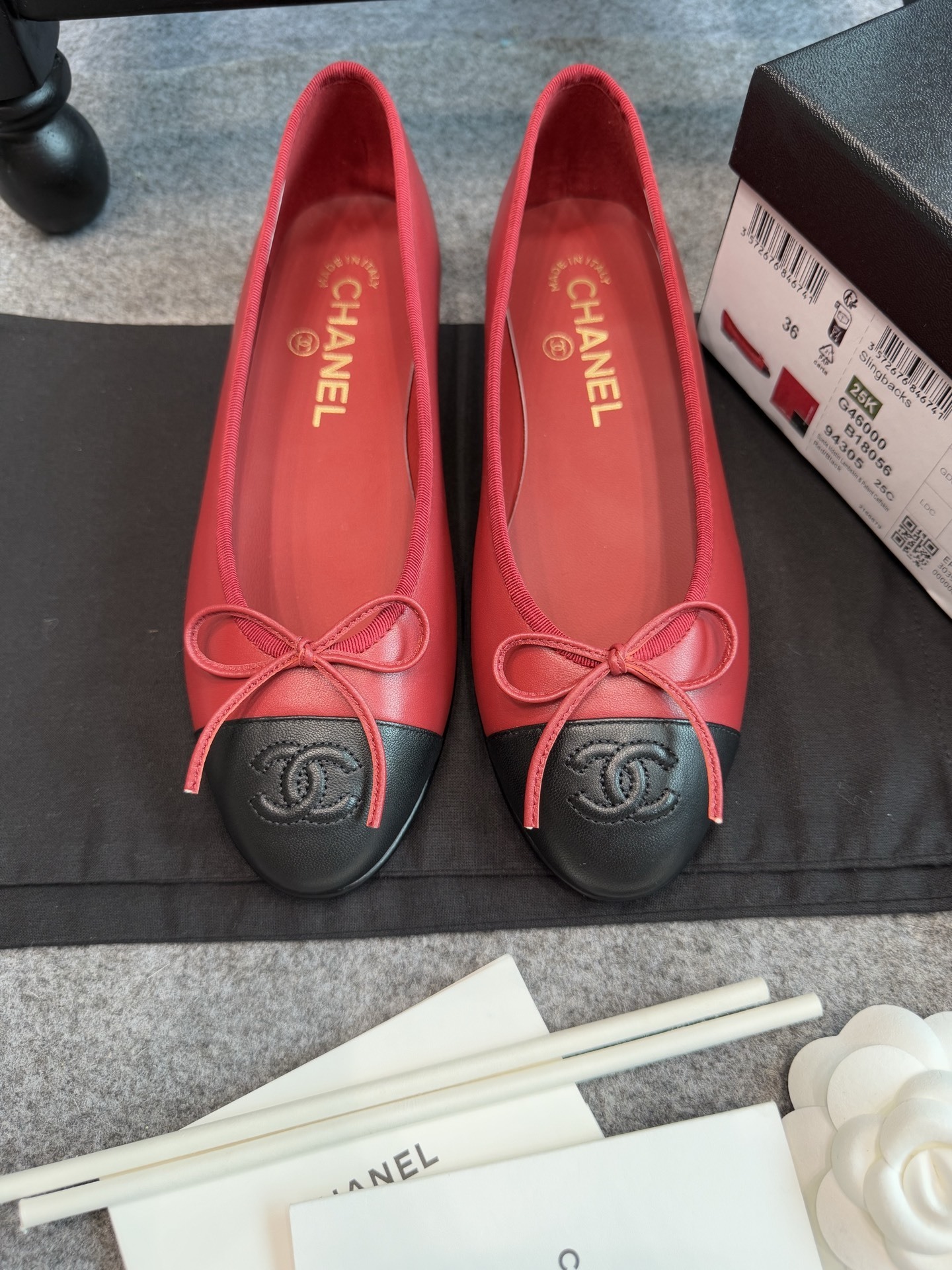 A pair of red Chanel ballet flats with black leather toe caps featuring the CC logo and a small bow, displayed with original packaging. - Hình 9