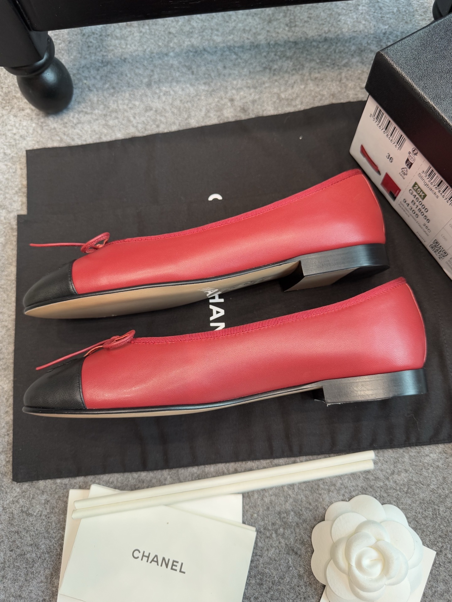 A pair of red Chanel ballet flats with black leather toe caps featuring the CC logo and a small bow, displayed with original packaging. - Hình 4