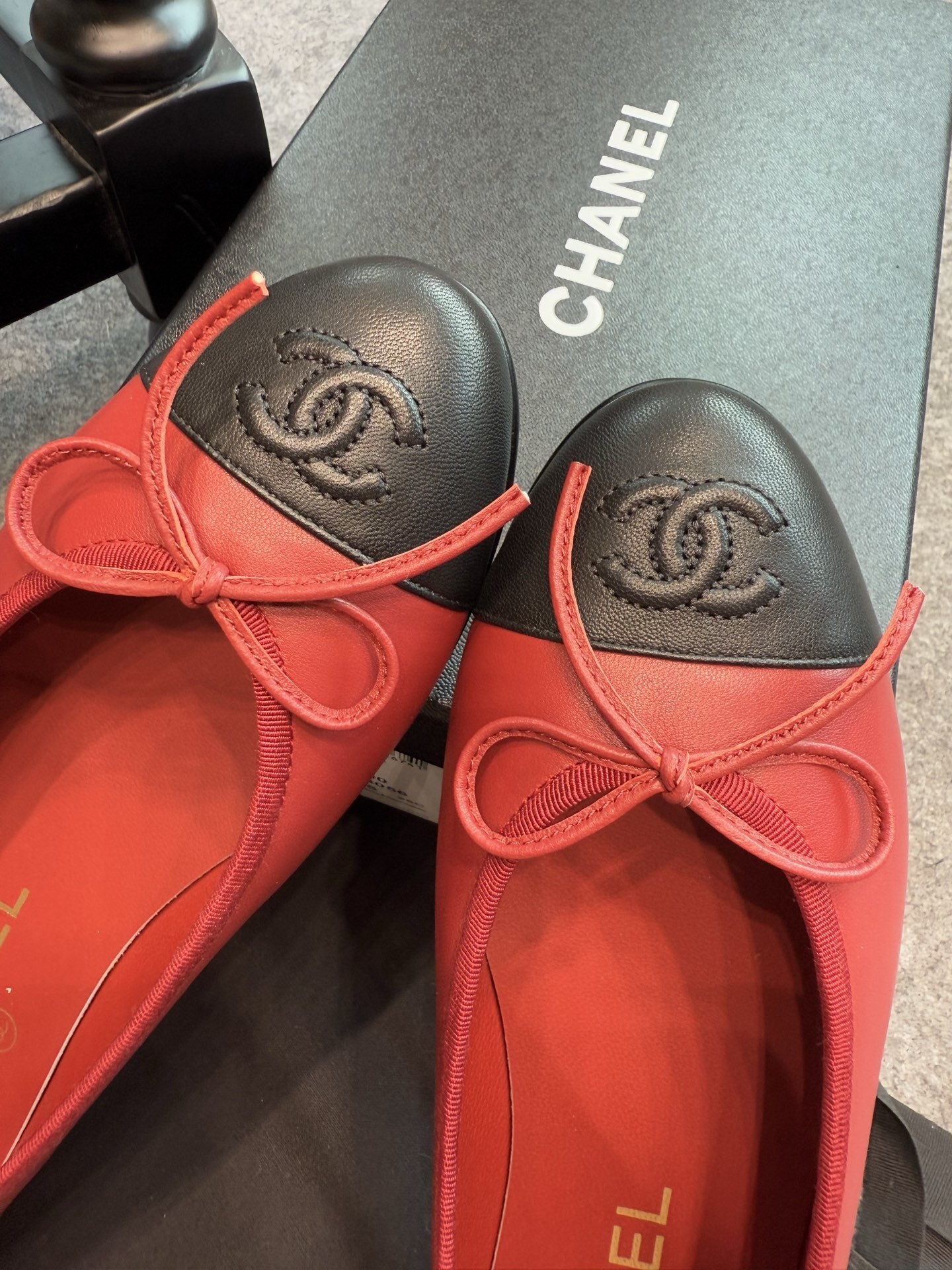 A pair of red Chanel ballet flats with black leather toe caps featuring the CC logo and a small bow, displayed with original packaging. - Hình 3