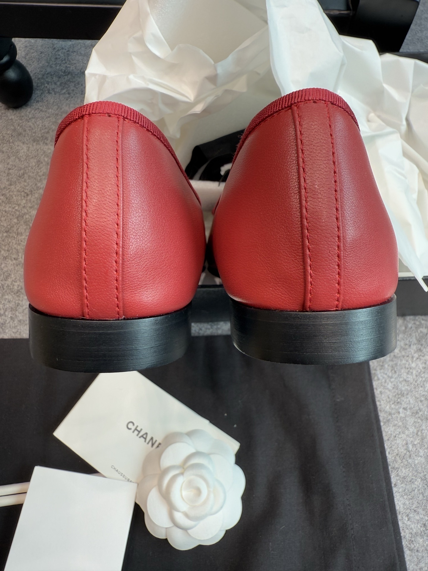 A pair of red Chanel ballet flats with black leather toe caps featuring the CC logo and a small bow, displayed with original packaging. - Hình 2