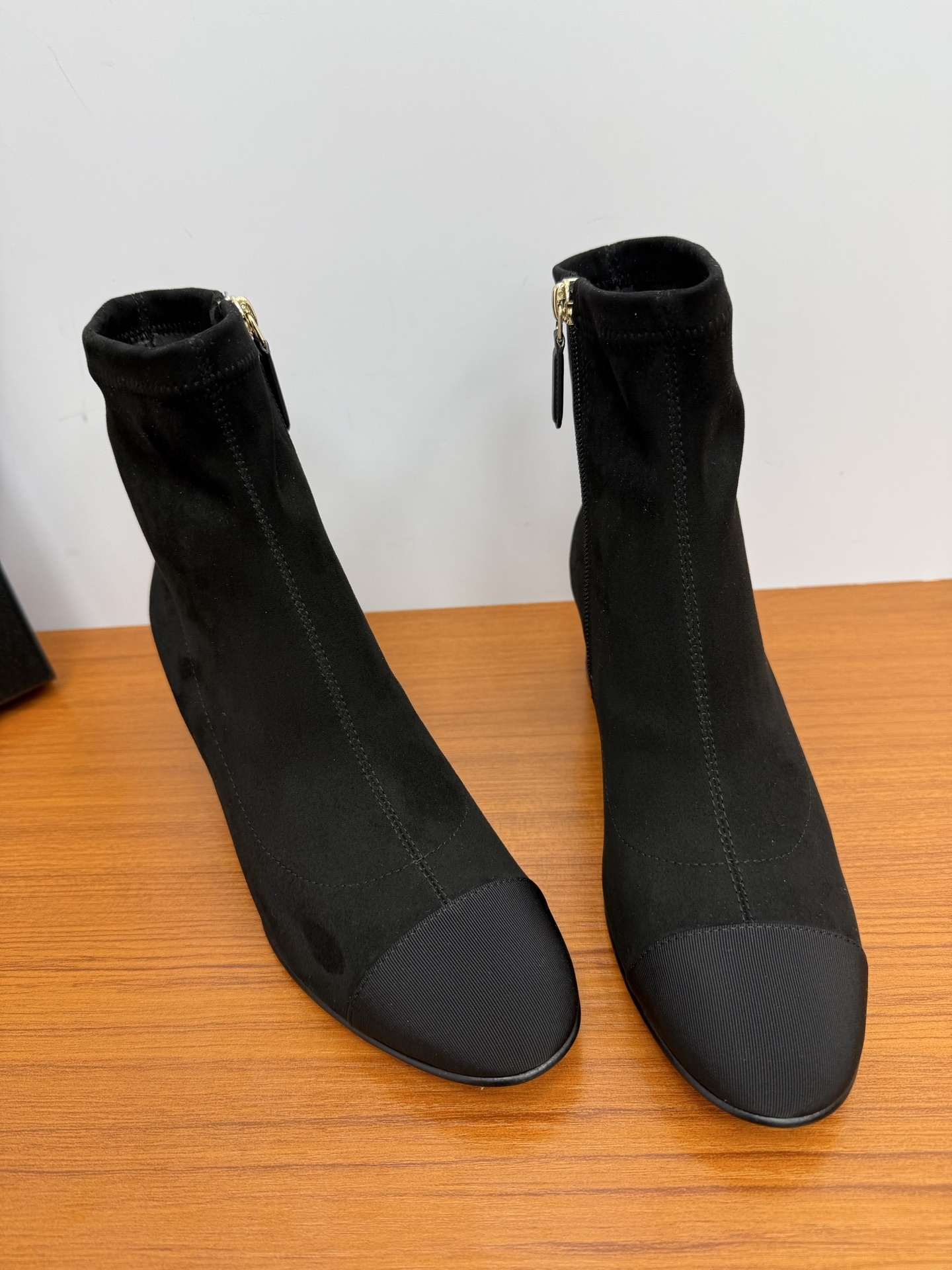A pair of sophisticated black suede ankle boots featuring a ribbed cap toe and gold side zippers on a wooden surface. - Hình 4