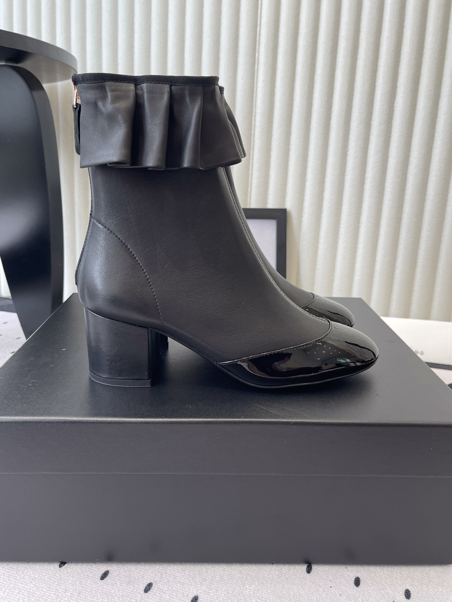 Luxury black leather ankle boots featuring a ruffle cuff detail and shiny patent leather cap toe on a designer box. - Hình 2