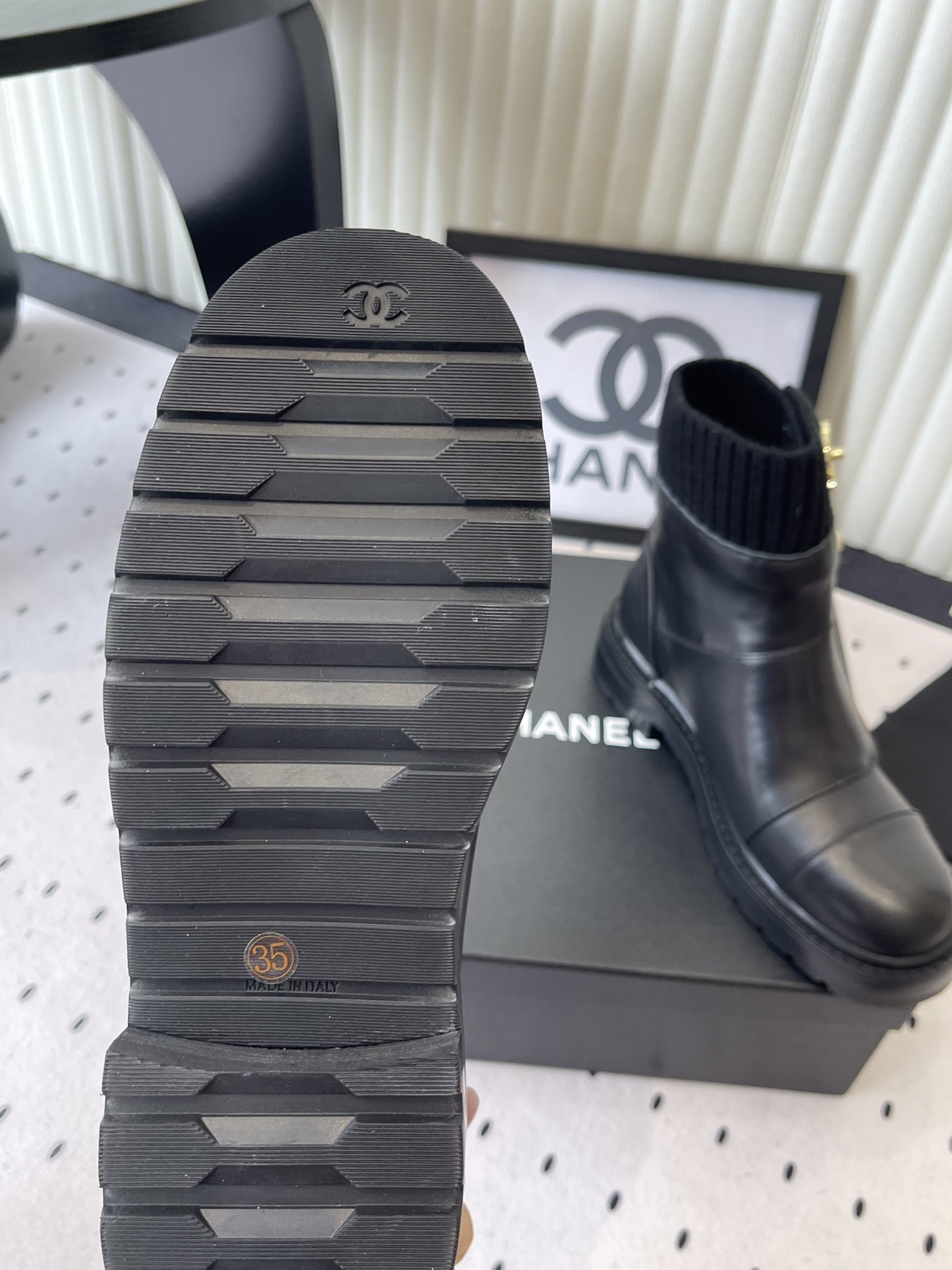 A pair of black leather ankle boots with ribbed knit collars and decorative gold CC logo hardware displayed on a black box. - Hình 9