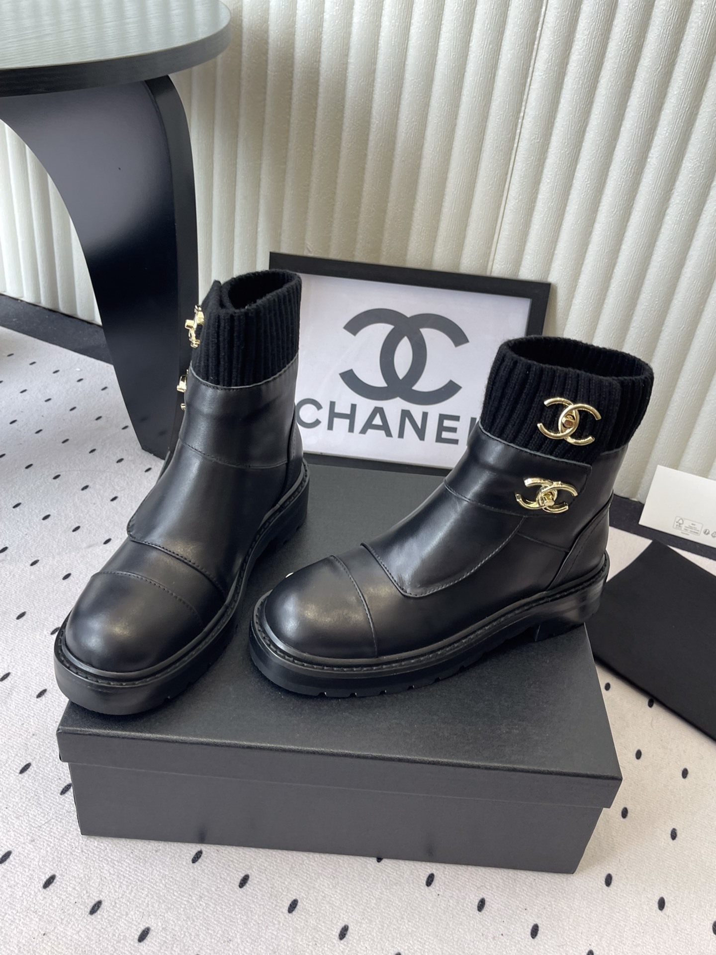 A pair of black leather ankle boots with ribbed knit collars and decorative gold CC logo hardware displayed on a black box. - Hình 8