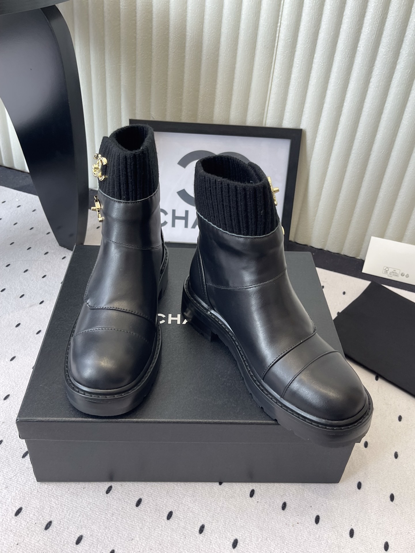 A pair of black leather ankle boots with ribbed knit collars and decorative gold CC logo hardware displayed on a black box. - Hình 7