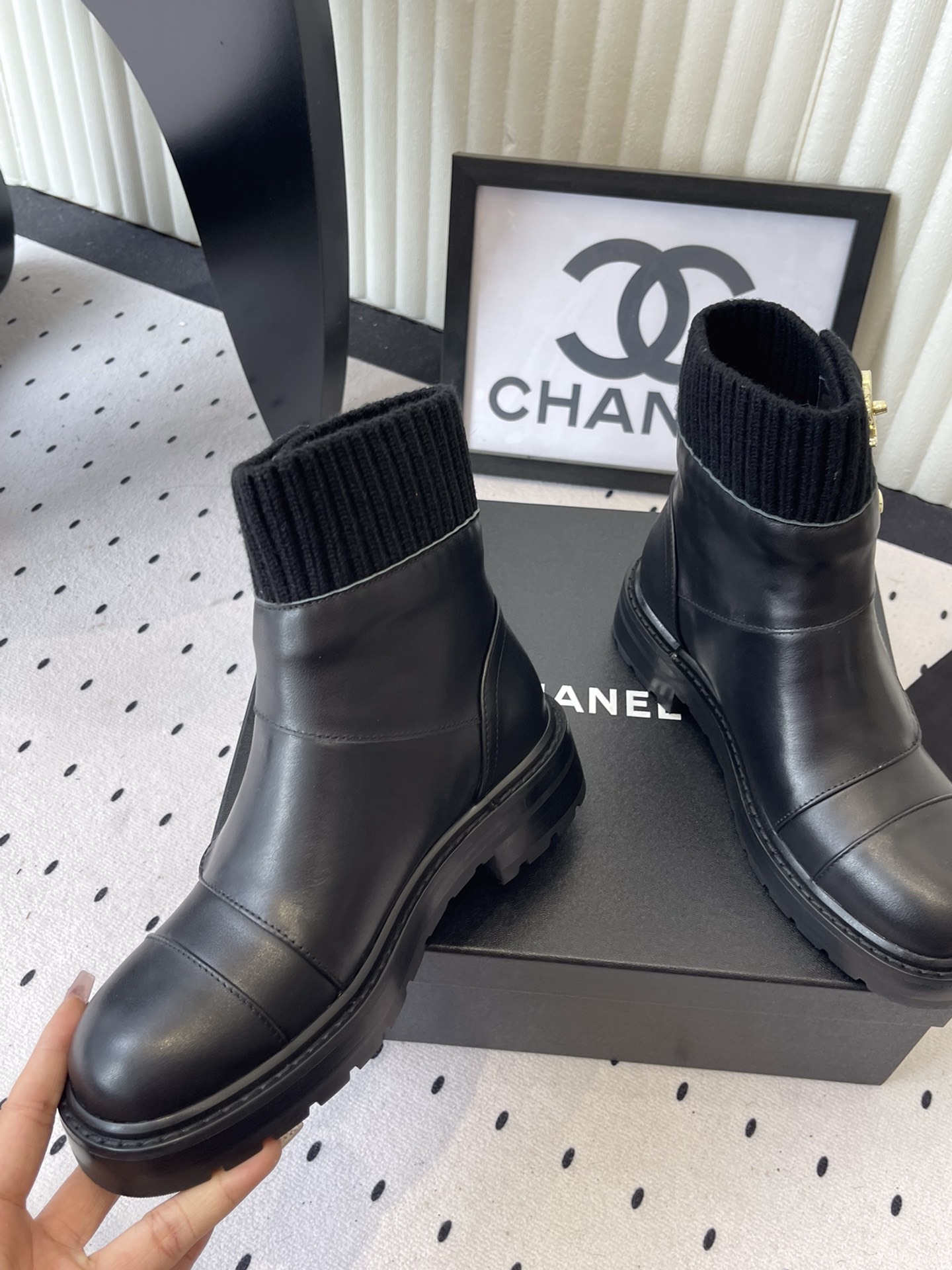 A pair of black leather ankle boots with ribbed knit collars and decorative gold CC logo hardware displayed on a black box. - Hình 5