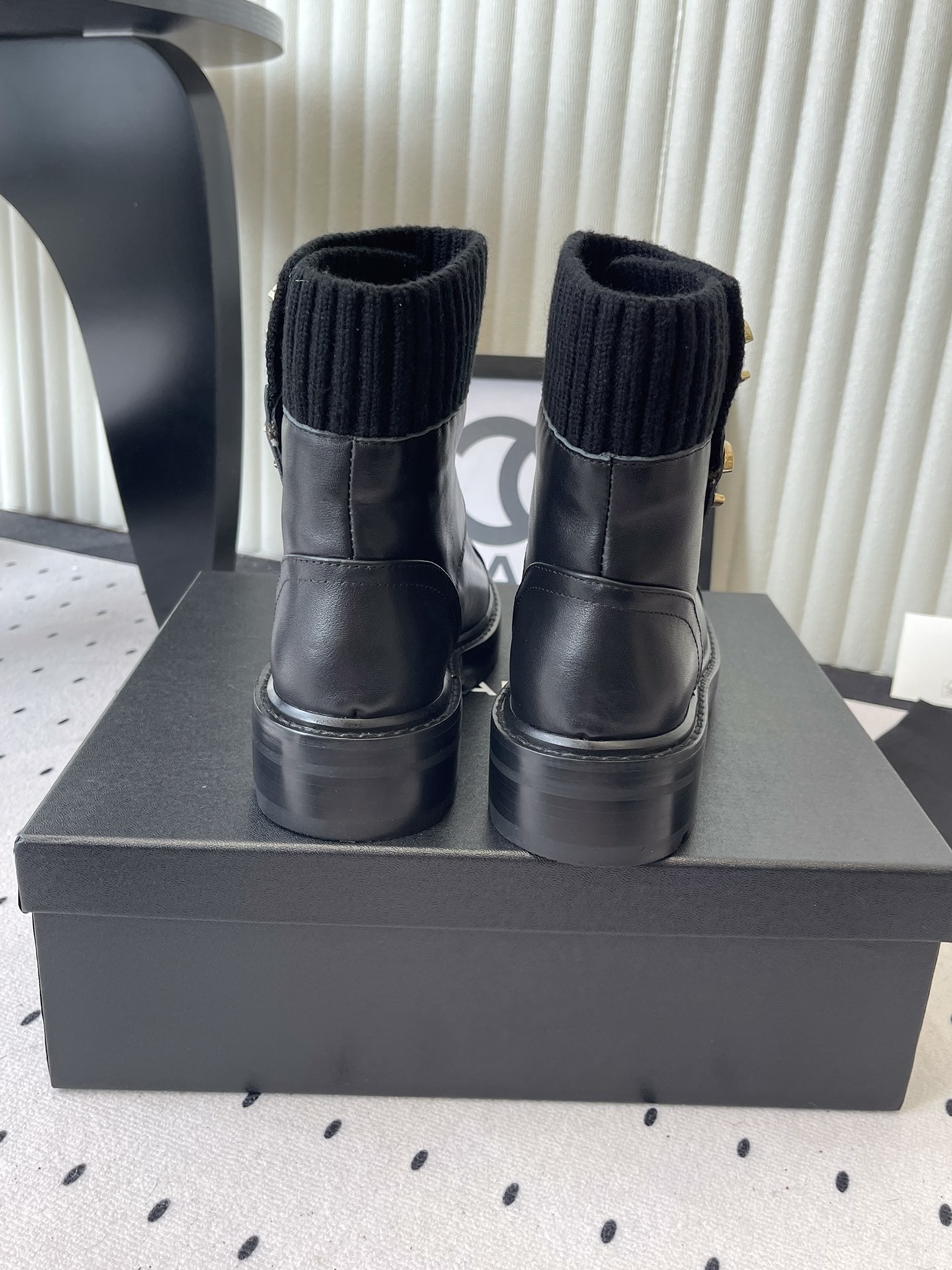 A pair of black leather ankle boots with ribbed knit collars and decorative gold CC logo hardware displayed on a black box. - Hình 3