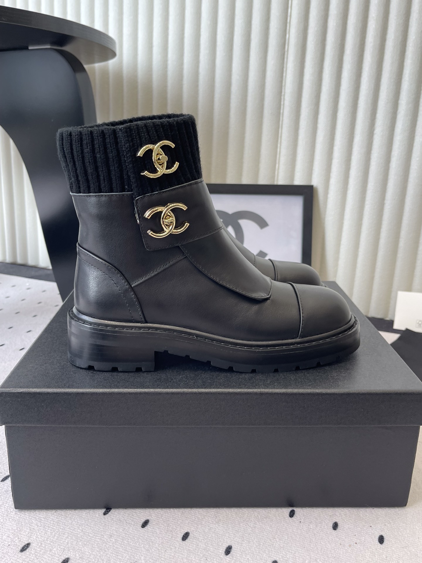 A pair of black leather ankle boots with ribbed knit collars and decorative gold CC logo hardware displayed on a black box. - Hình 2