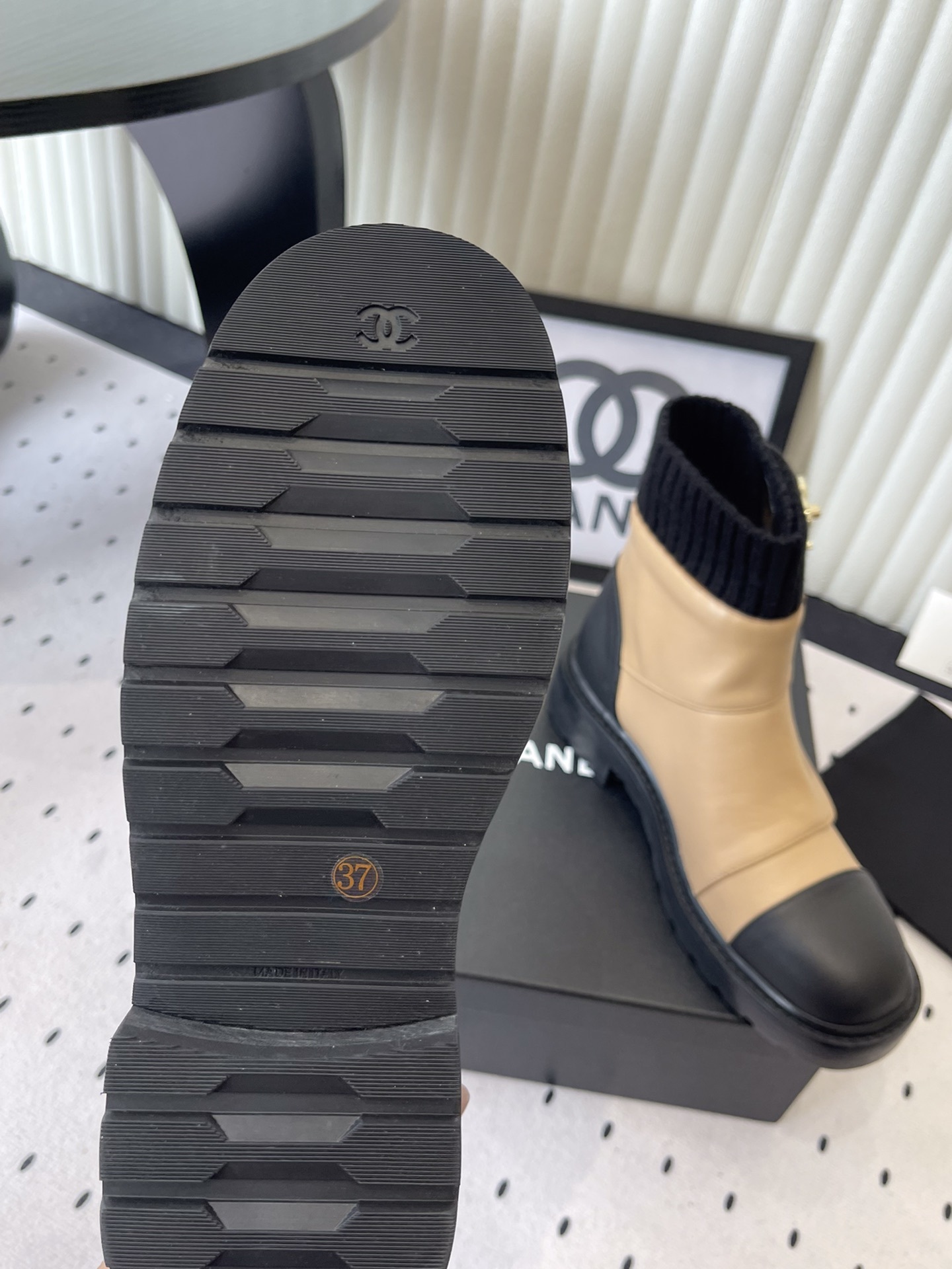 Pair of Chanel beige and black leather ankle boots with gold CC logo and black knit sock collar displayed on a black box. - Hình 9
