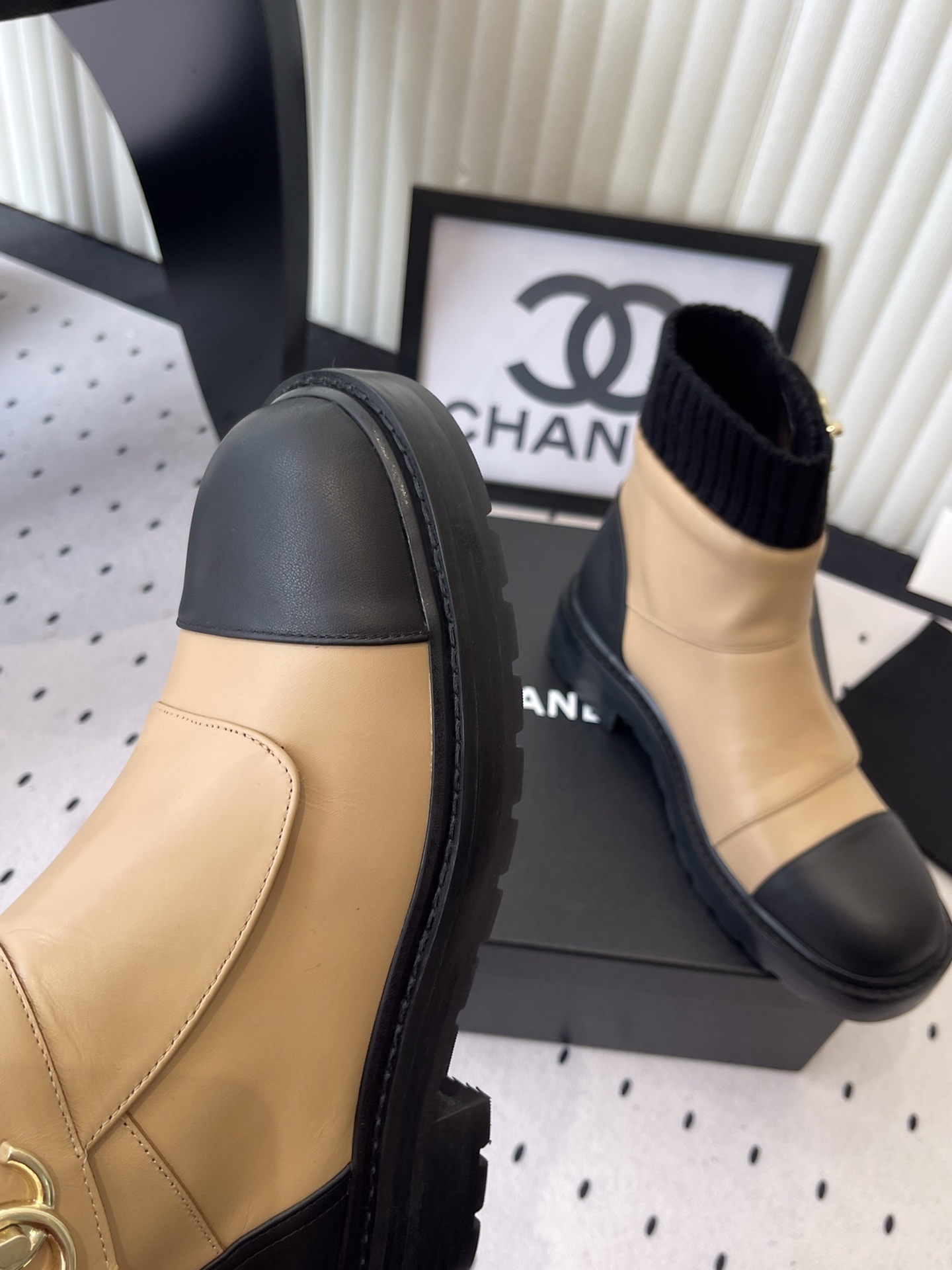 Pair of Chanel beige and black leather ankle boots with gold CC logo and black knit sock collar displayed on a black box. - Hình 6