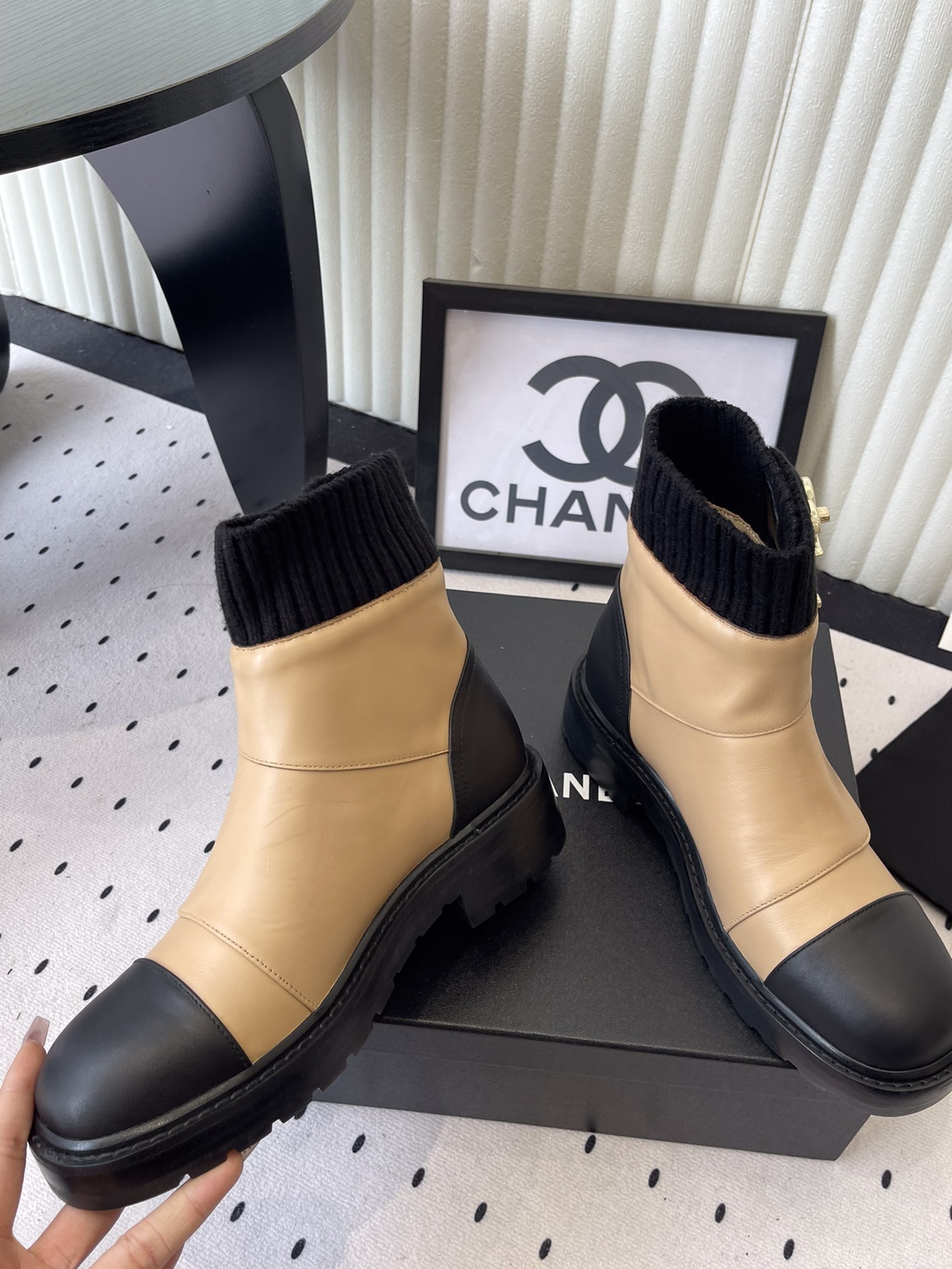 Pair of Chanel beige and black leather ankle boots with gold CC logo and black knit sock collar displayed on a black box. - Hình 5