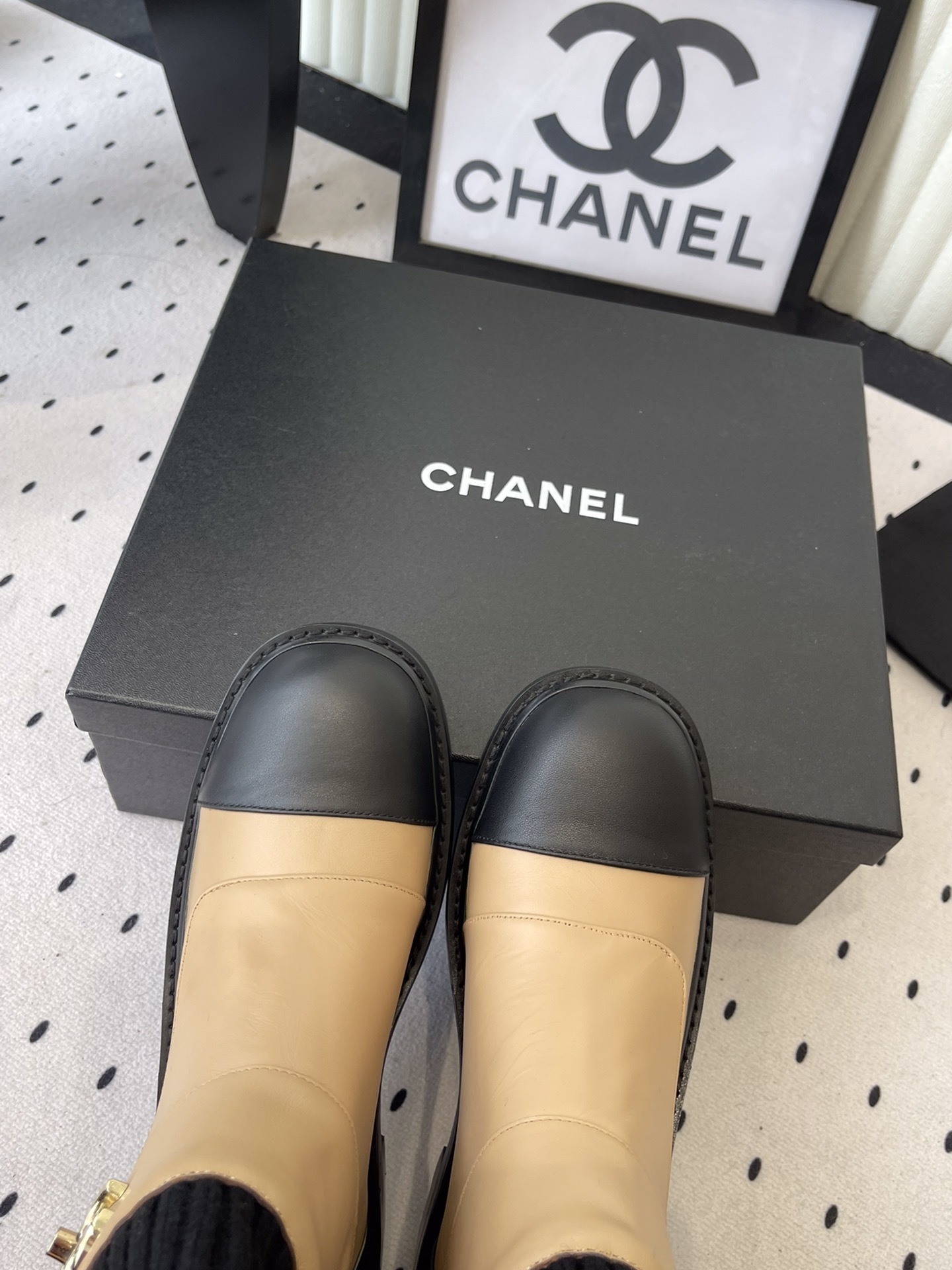 Pair of Chanel beige and black leather ankle boots with gold CC logo and black knit sock collar displayed on a black box. - Hình 4