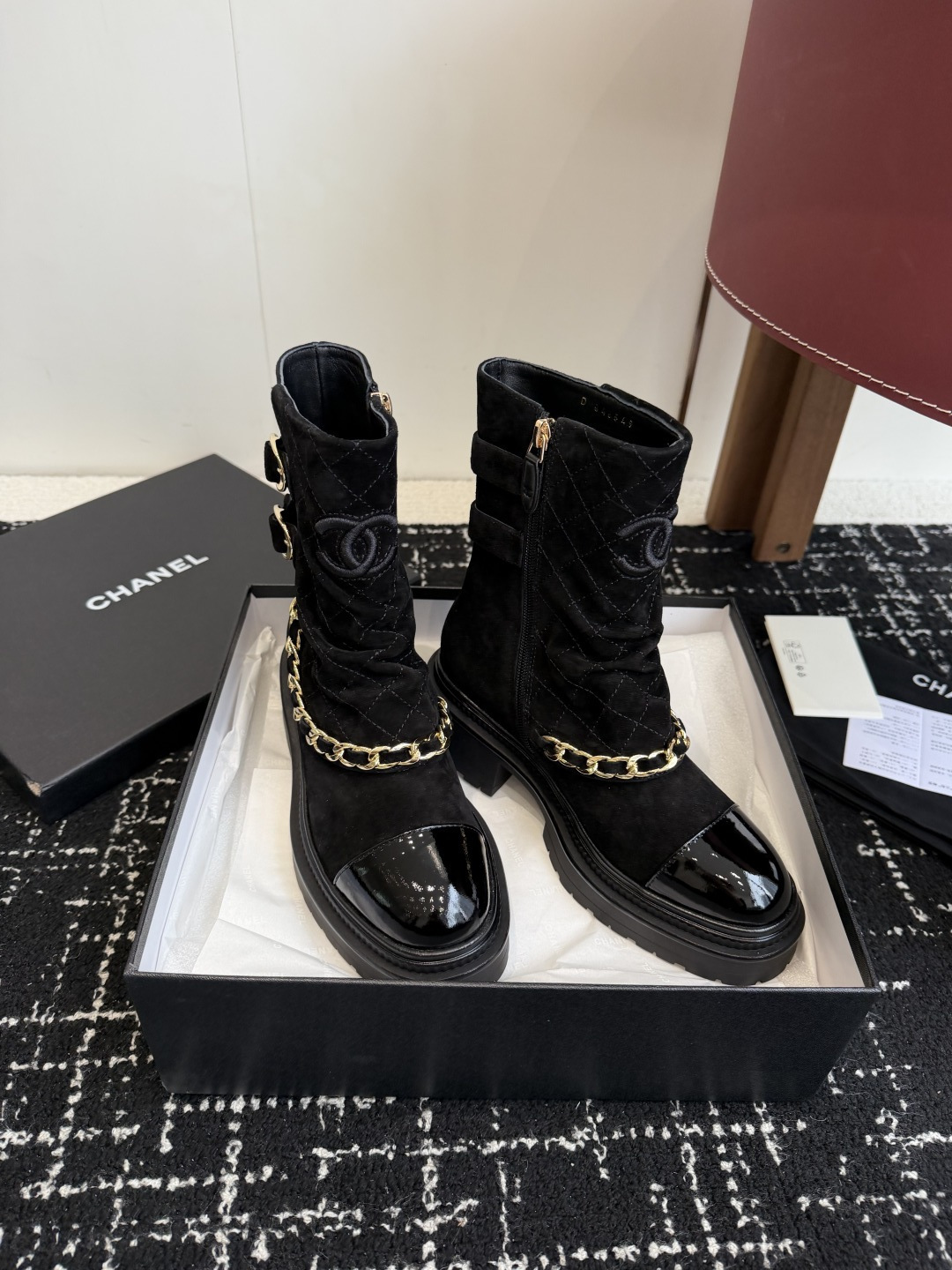 Pair of black Chanel quilted suede ankle boots with gold chain and patent leather toe caps on a black box - Hình 3