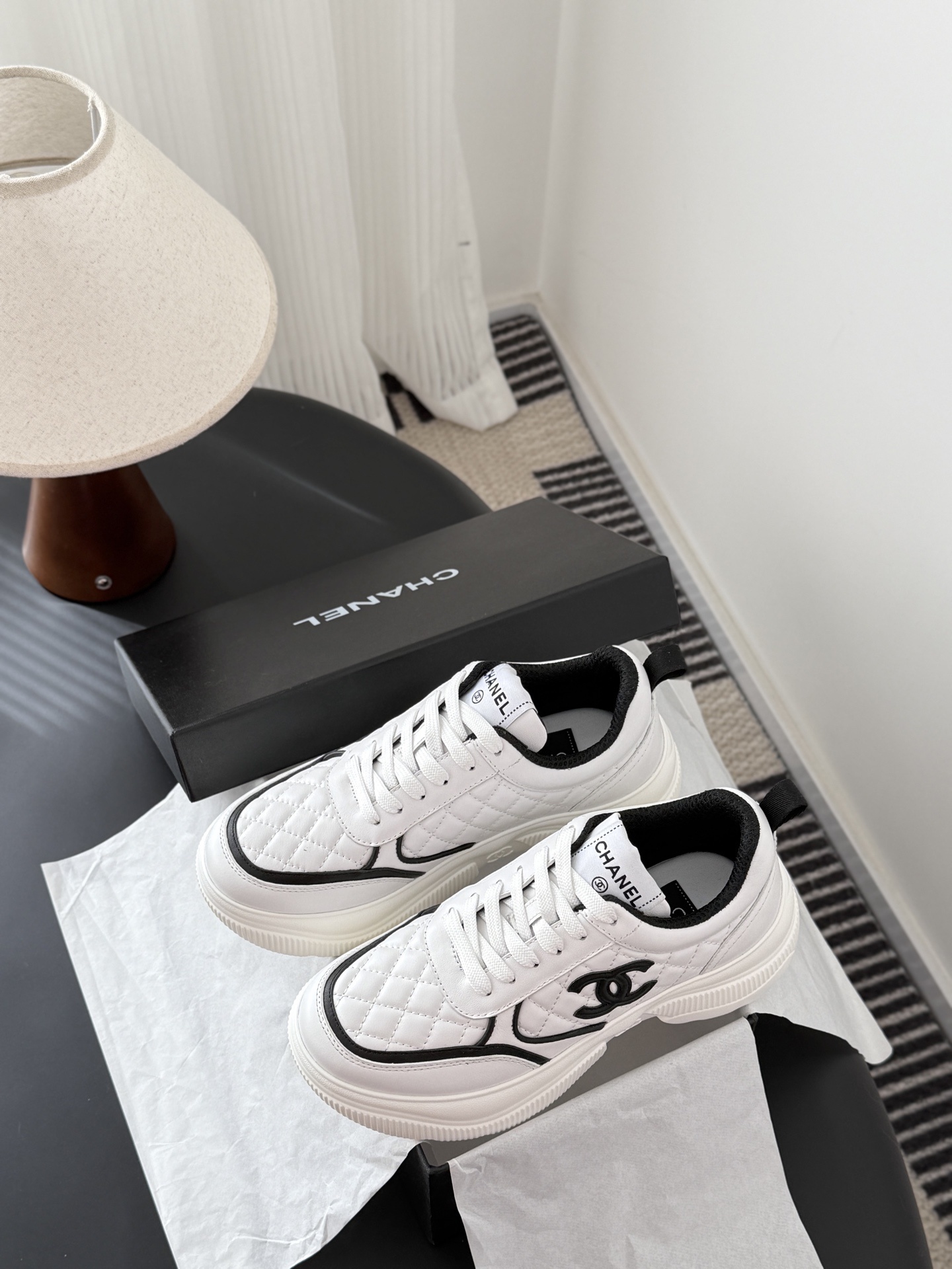 A pair of white Chanel quilted leather sneakers with black CC logo and chunky white soles next to a black Chanel box - Hình 9
