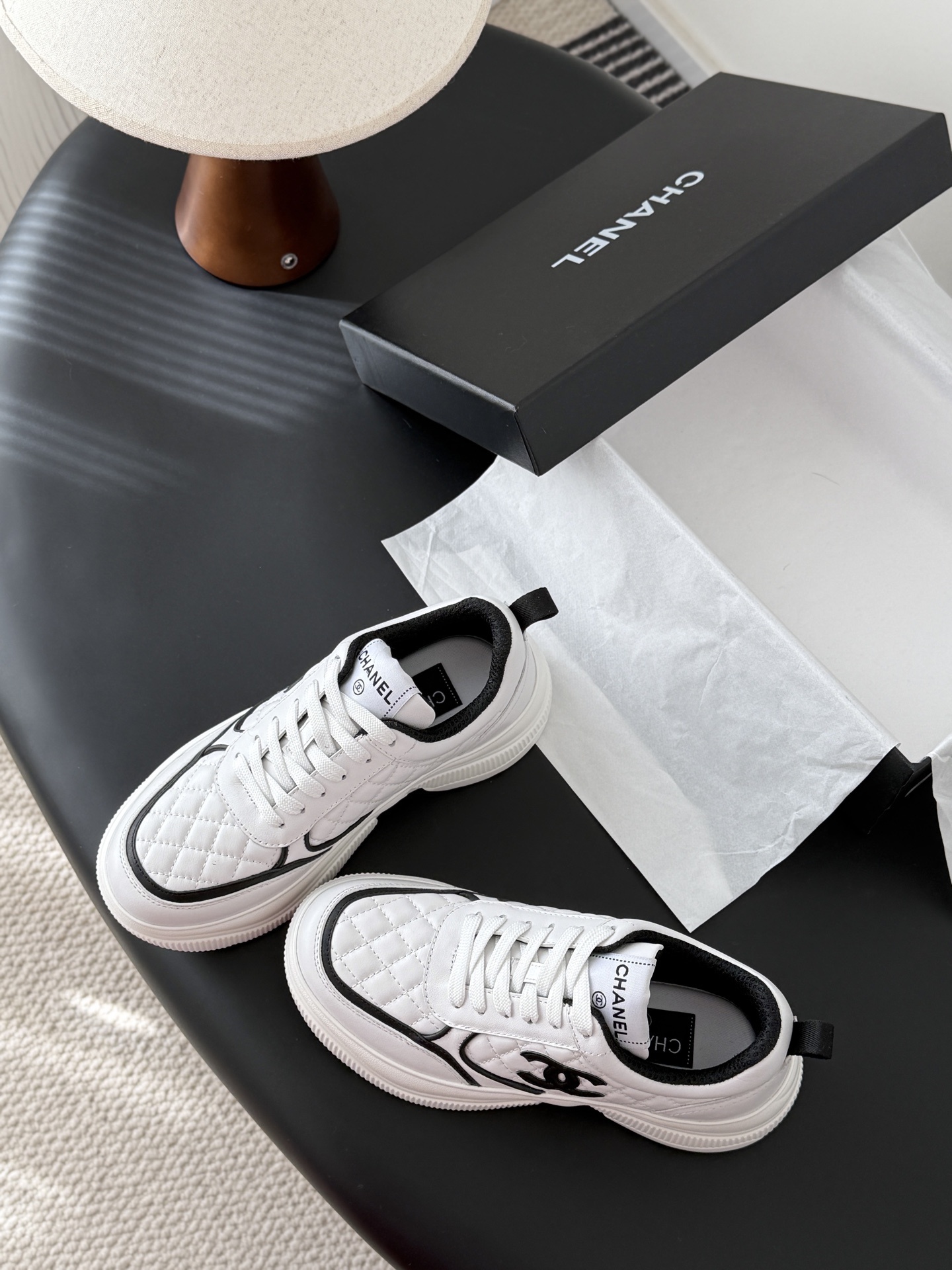 A pair of white Chanel quilted leather sneakers with black CC logo and chunky white soles next to a black Chanel box - Hình 6