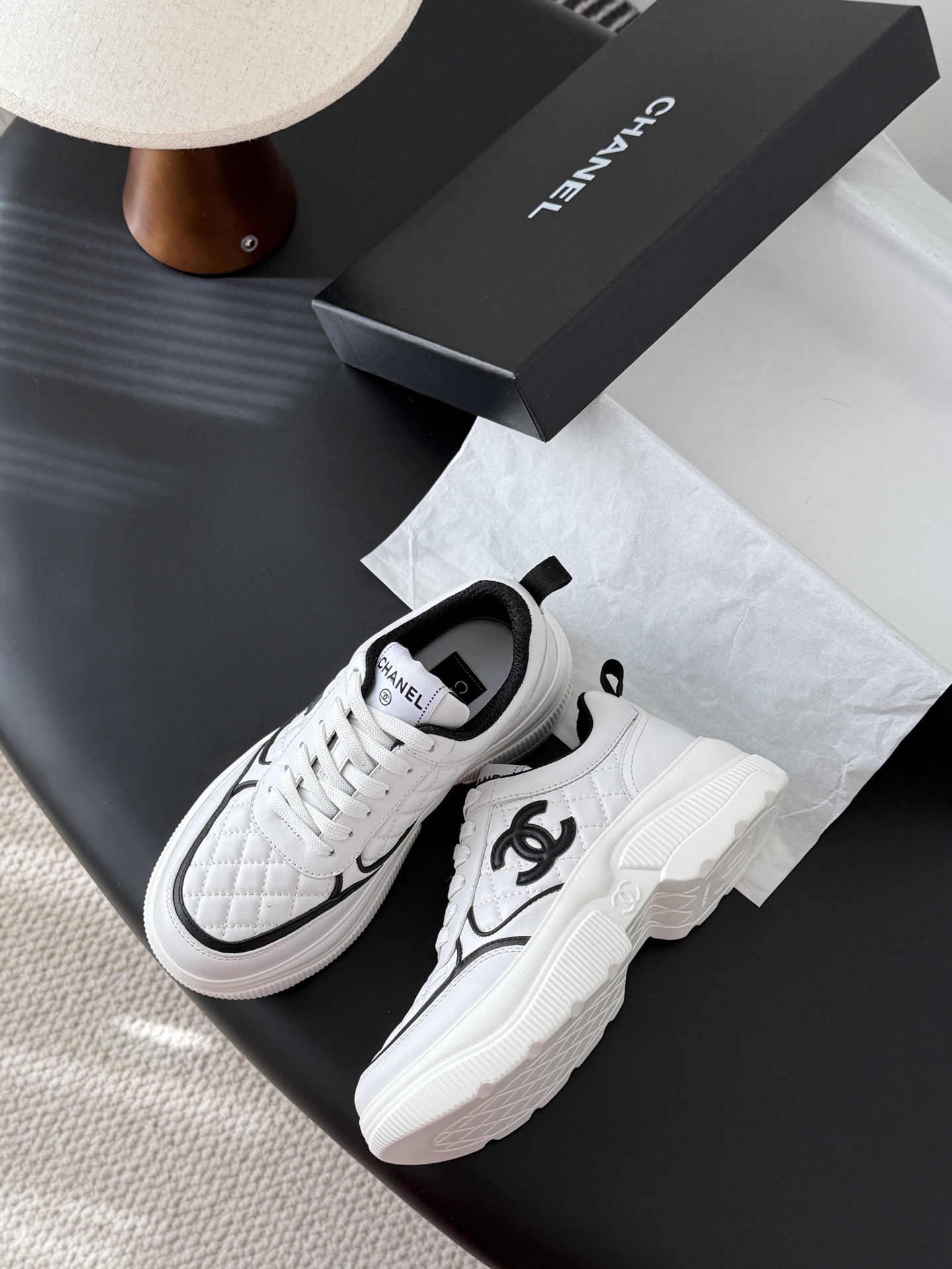 A pair of white Chanel quilted leather sneakers with black CC logo and chunky white soles next to a black Chanel box - Hình 4