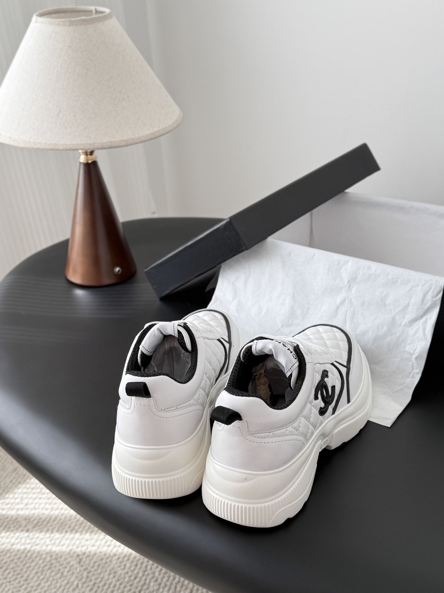 A pair of white Chanel quilted leather sneakers with black CC logo and chunky white soles next to a black Chanel box - Hình 3