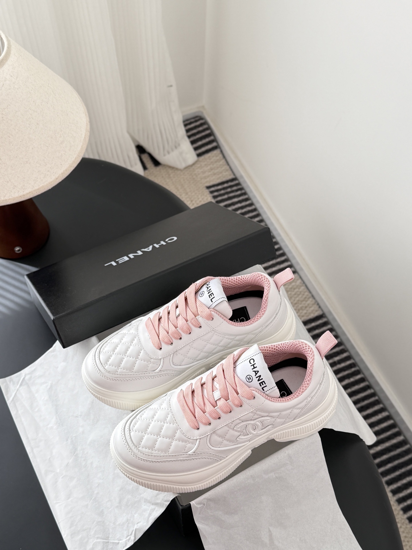 A pair of Chanel white quilted sneakers with pink laces and pink interior lining next to a black Chanel shoe box - Hình 9