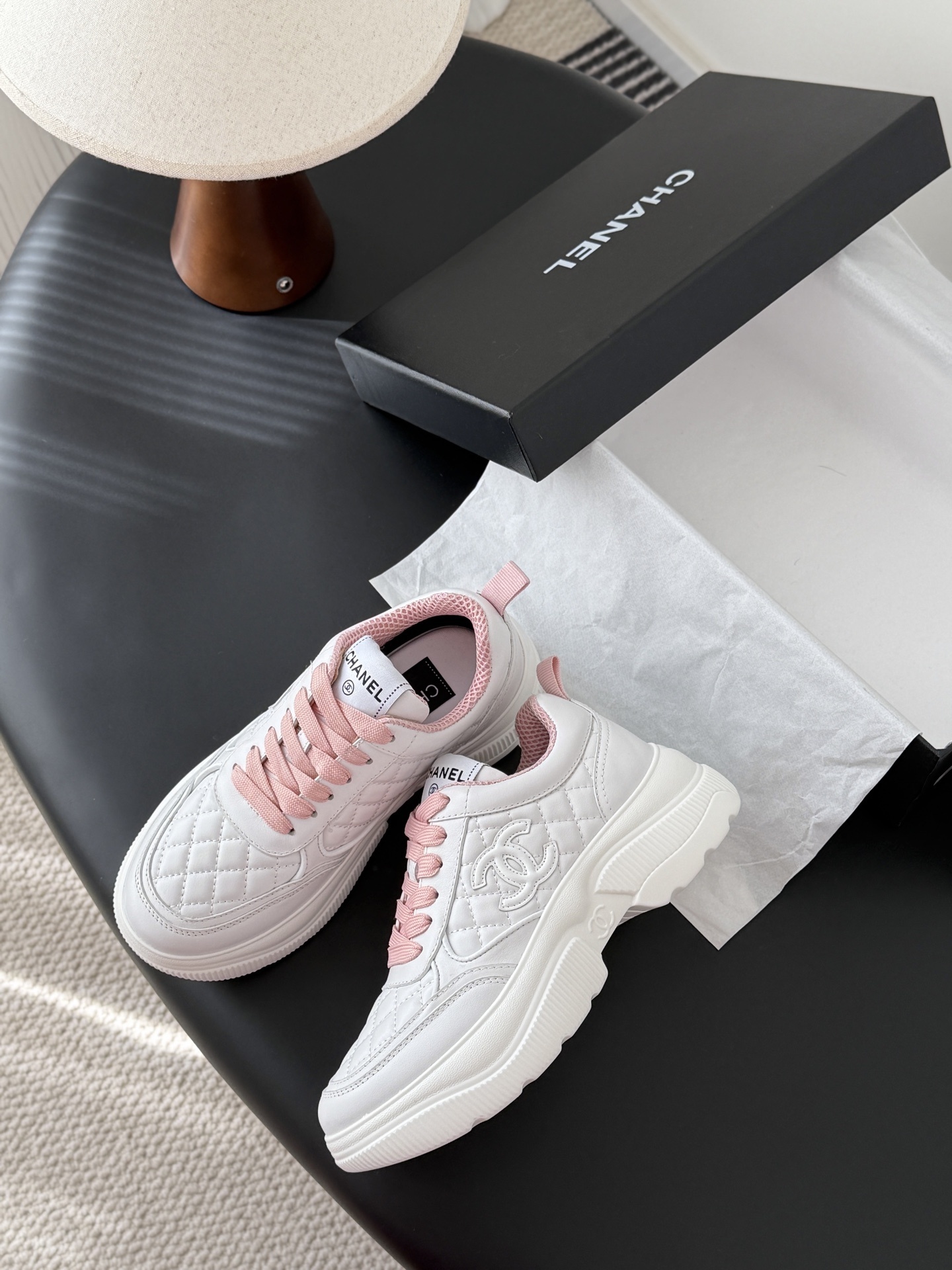 A pair of Chanel white quilted sneakers with pink laces and pink interior lining next to a black Chanel shoe box - Hình 4