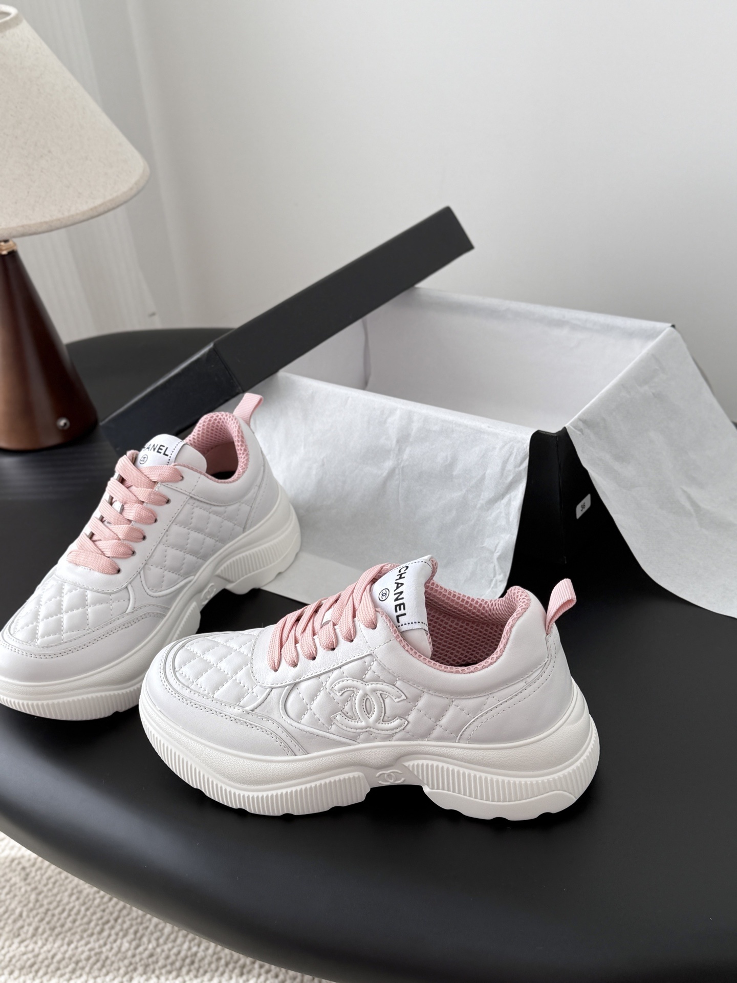 A pair of Chanel white quilted sneakers with pink laces and pink interior lining next to a black Chanel shoe box - Hình 2