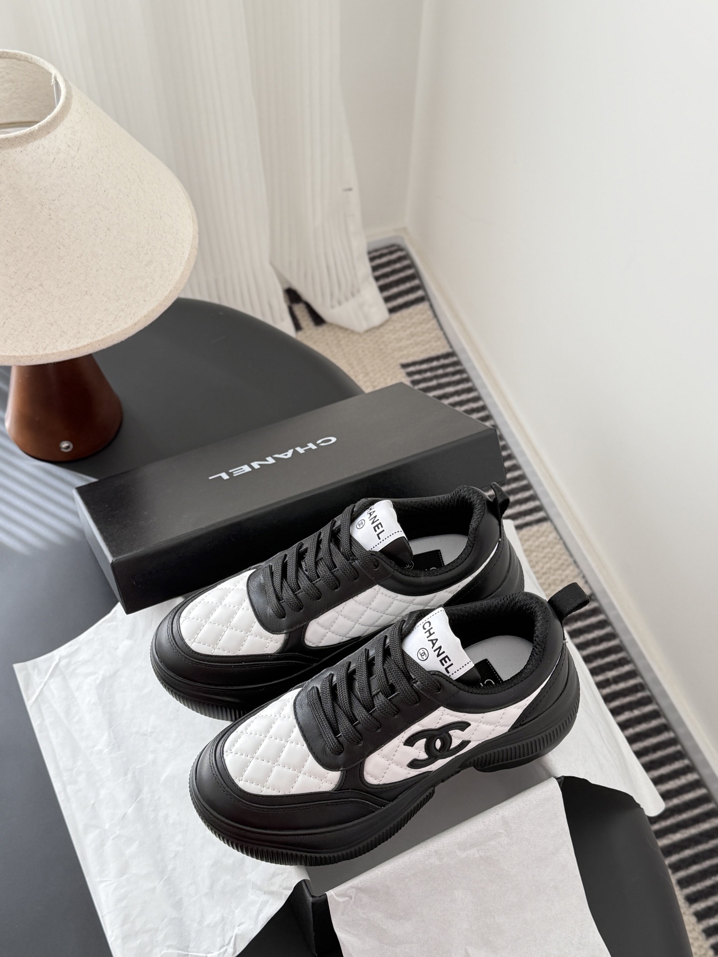 Pair of black and white Chanel quilted leather sneakers with CC logo on a black table next to a branded box - Hình 9