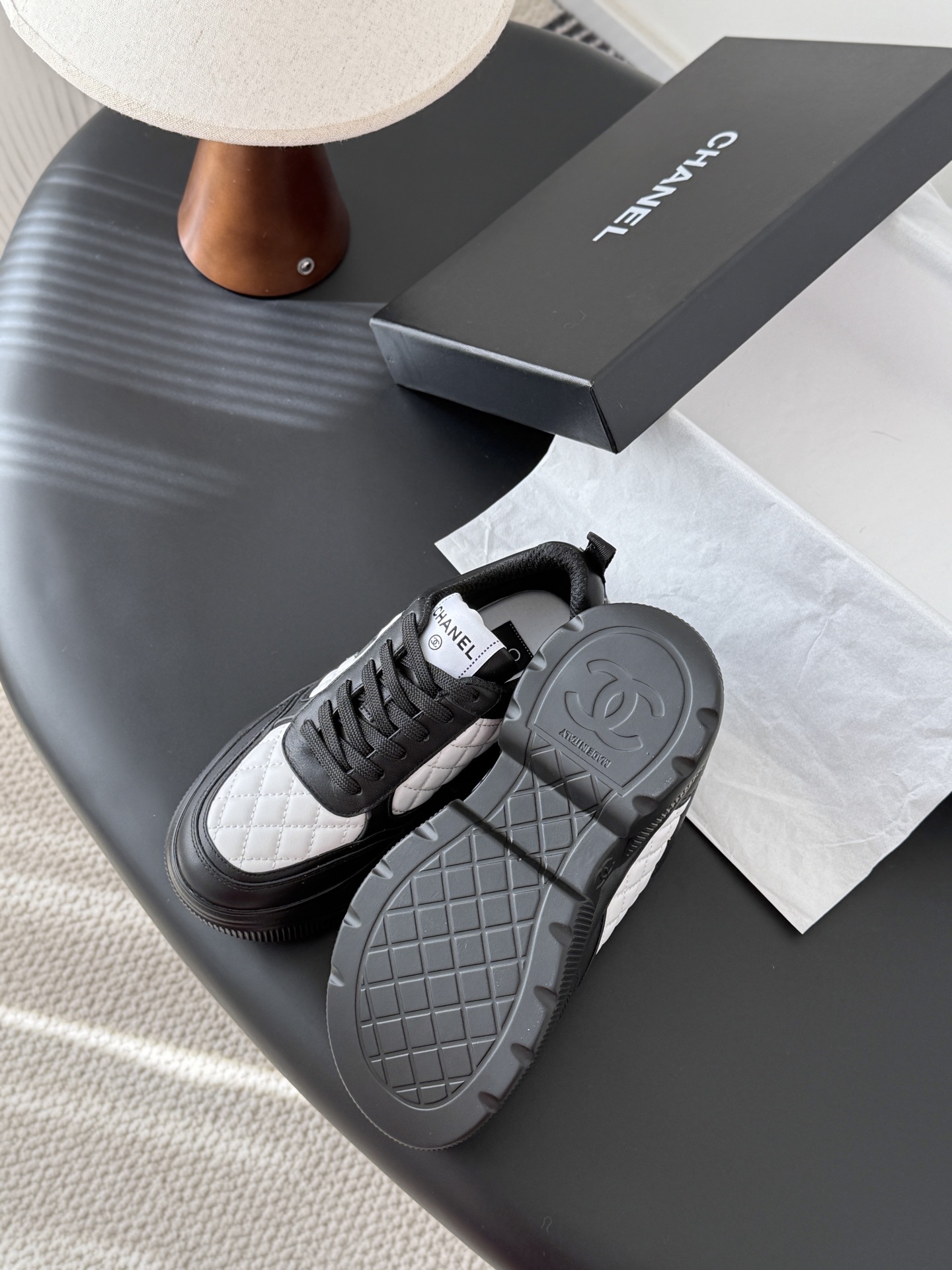 Pair of black and white Chanel quilted leather sneakers with CC logo on a black table next to a branded box - Hình 5