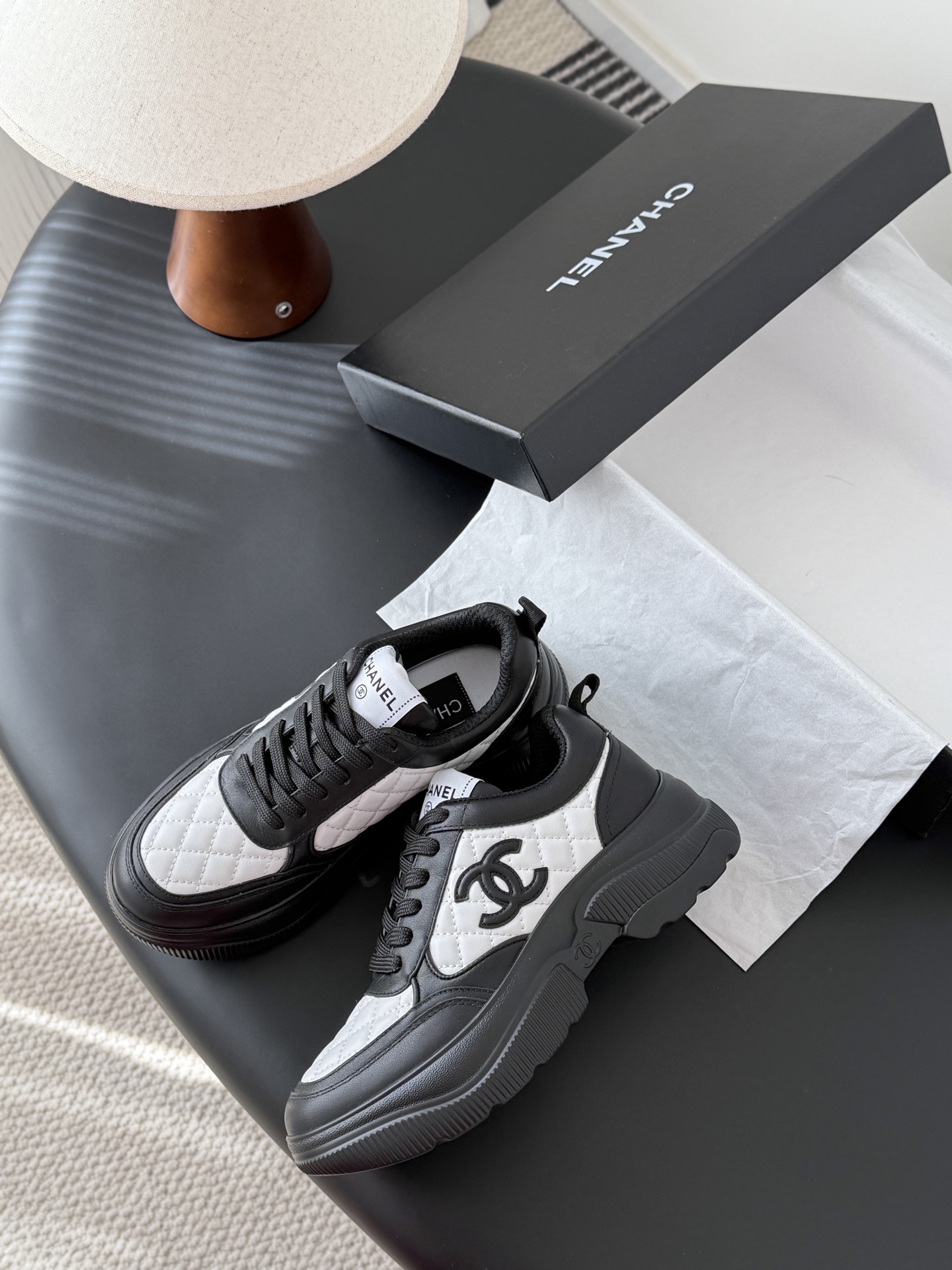 Pair of black and white Chanel quilted leather sneakers with CC logo on a black table next to a branded box - Hình 4