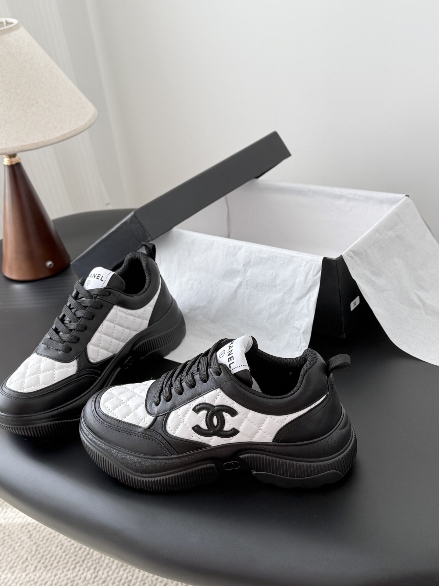 Pair of black and white Chanel quilted leather sneakers with CC logo on a black table next to a branded box - Hình 2