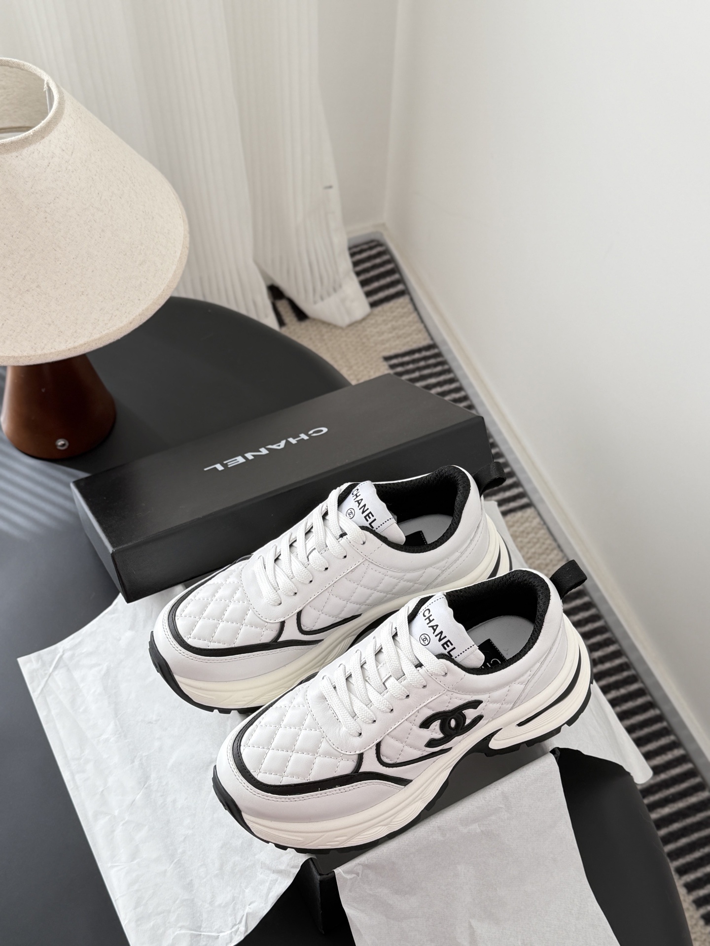 A pair of white Chanel chunky sneakers with black quilted details and CC logo displayed on a table next to a black Chanel box. - Hình 9