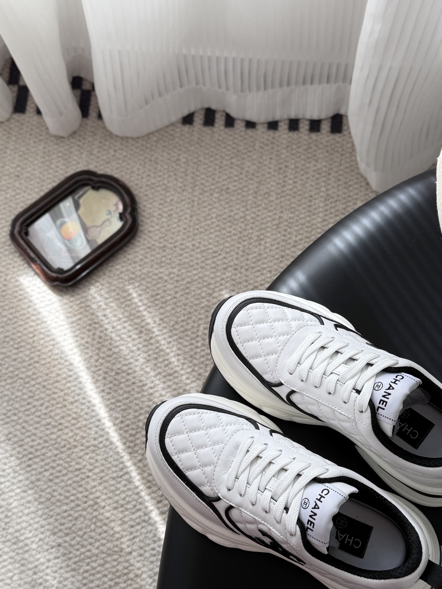 A pair of white Chanel chunky sneakers with black quilted details and CC logo displayed on a table next to a black Chanel box. - Hình 7