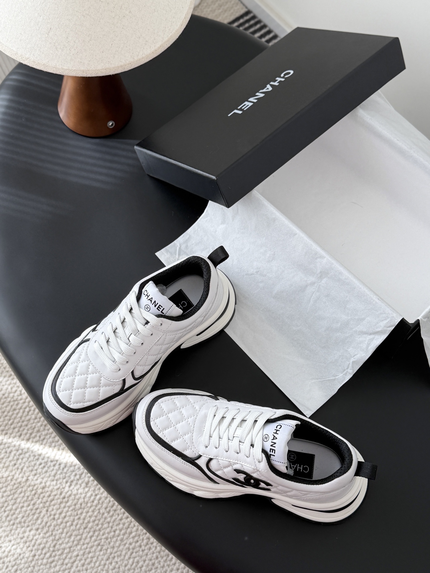 A pair of white Chanel chunky sneakers with black quilted details and CC logo displayed on a table next to a black Chanel box. - Hình 6