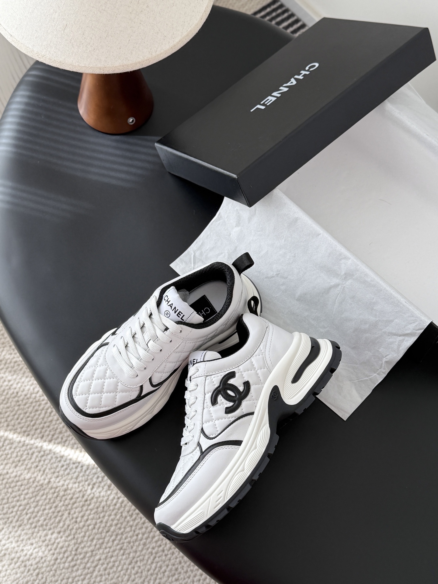 A pair of white Chanel chunky sneakers with black quilted details and CC logo displayed on a table next to a black Chanel box. - Hình 4