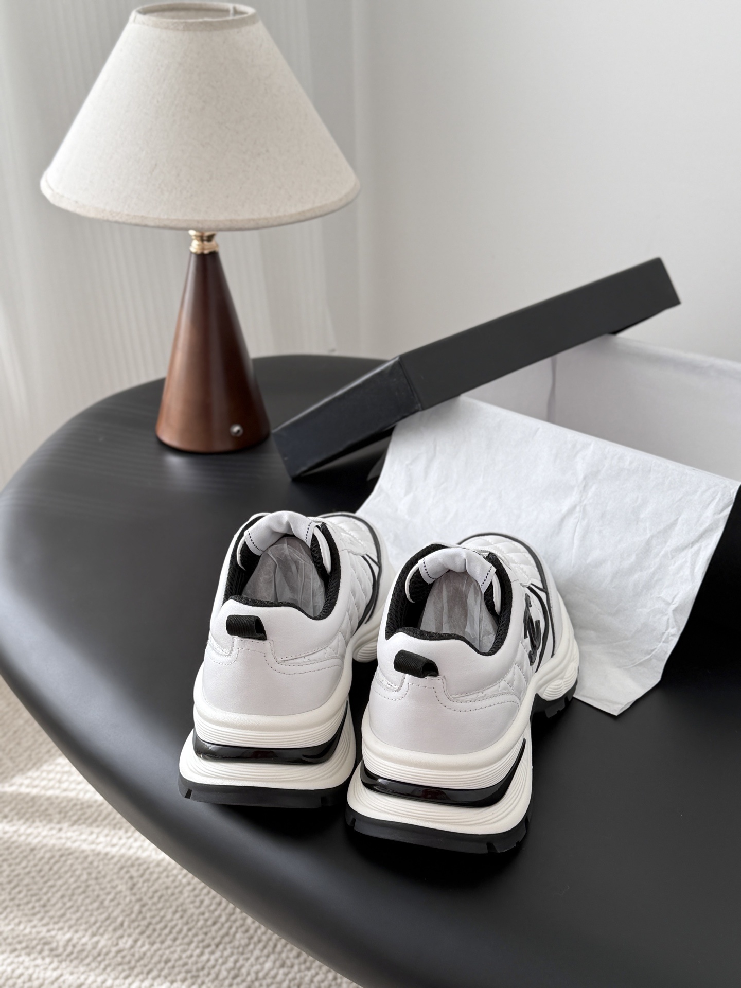 A pair of white Chanel chunky sneakers with black quilted details and CC logo displayed on a table next to a black Chanel box. - Hình 3