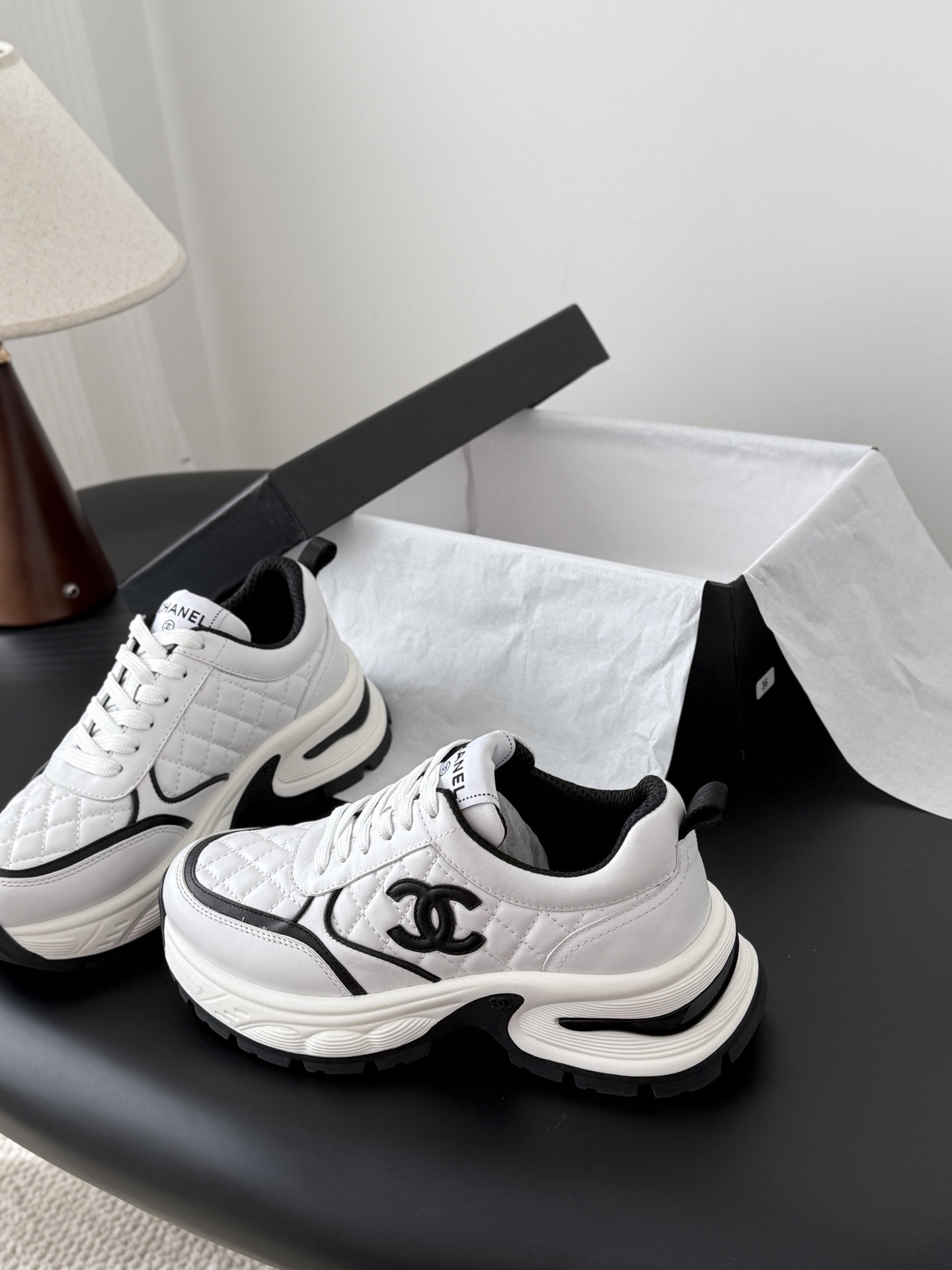 A pair of white Chanel chunky sneakers with black quilted details and CC logo displayed on a table next to a black Chanel box. - Hình 2