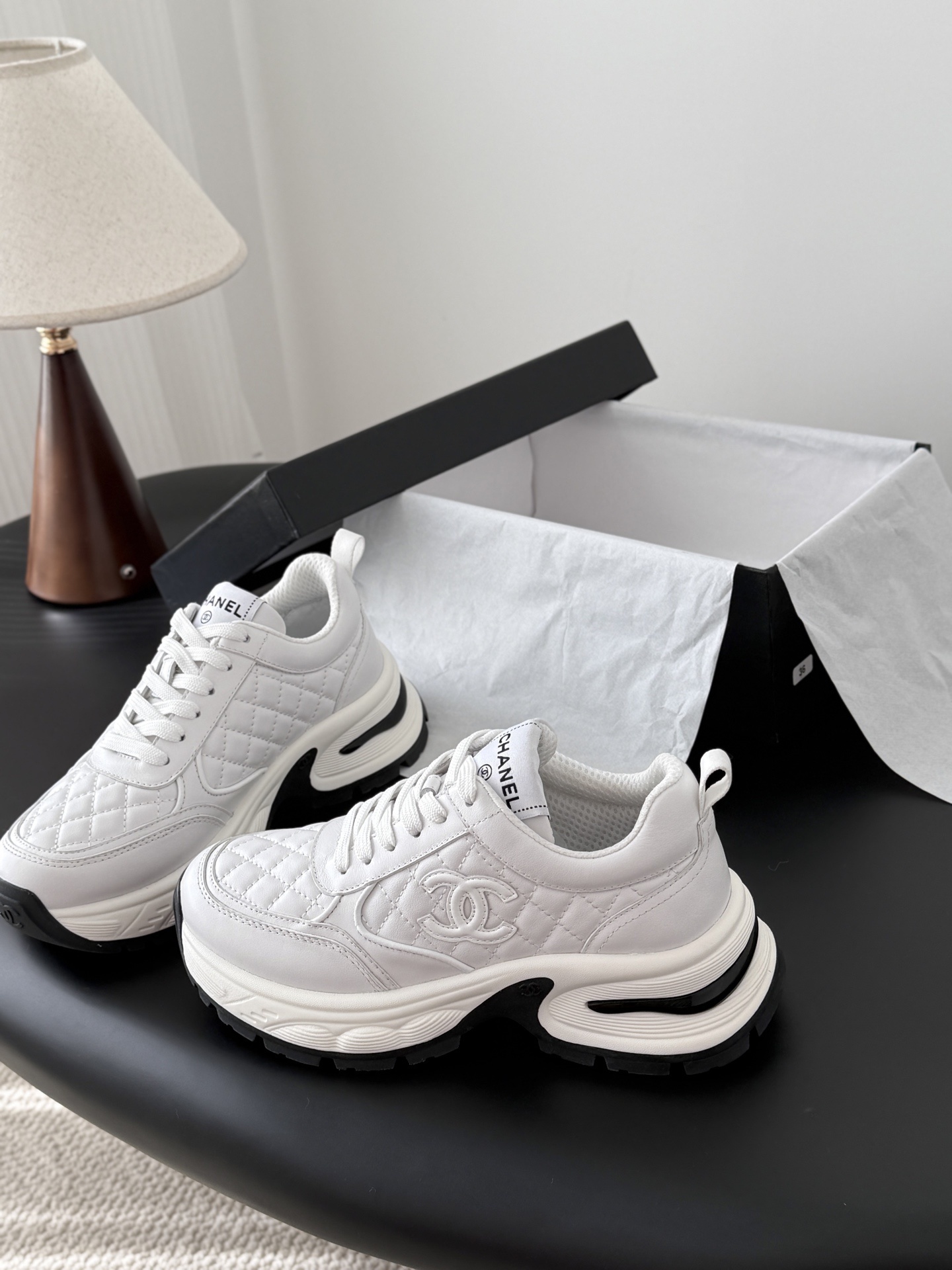 A pair of luxury white Chanel quilted leather sneakers with chunky soles and CC logo on a black table - Hình 2