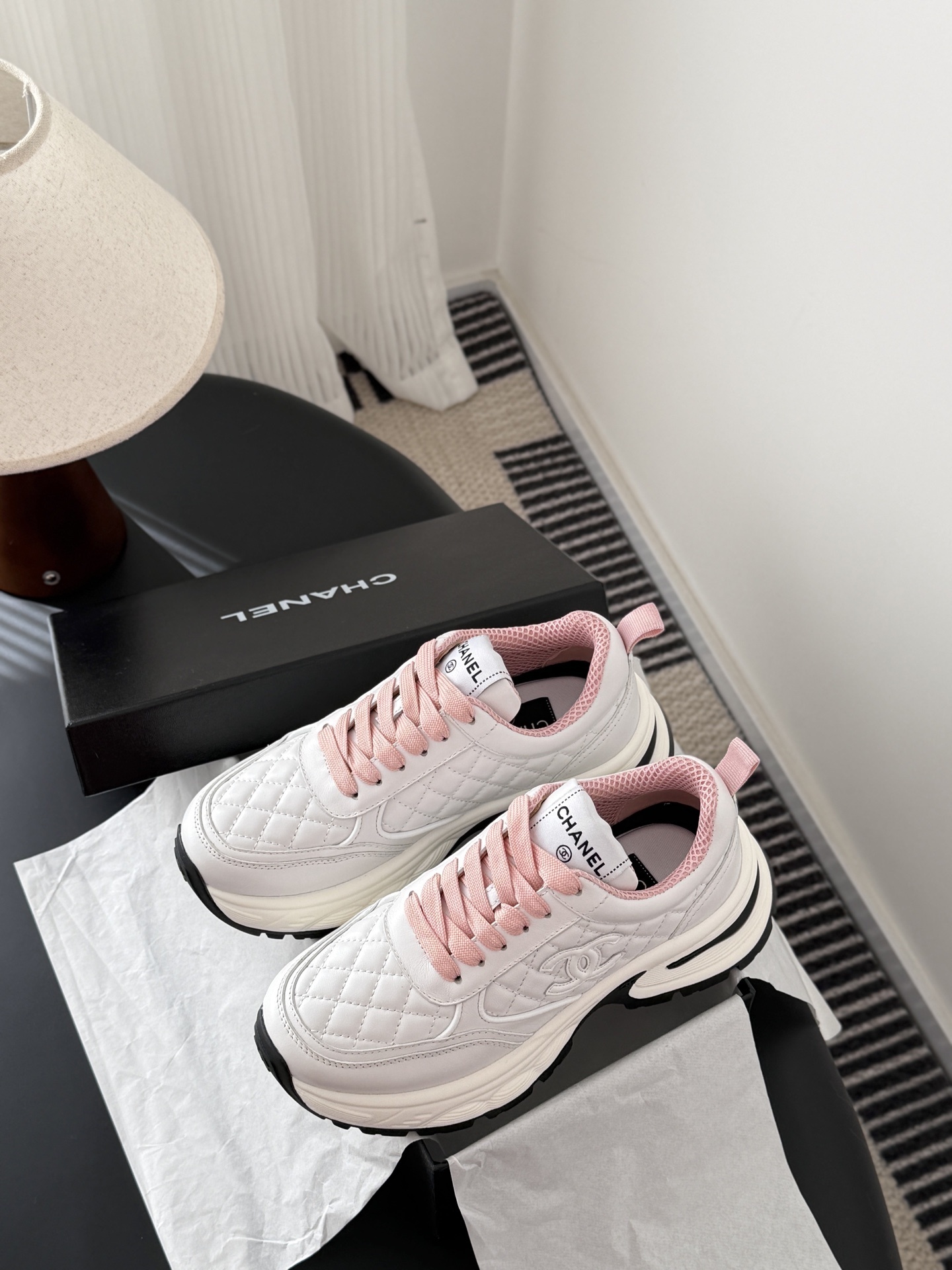 A pair of luxury white Chanel sneakers with pink laces and quilted leather detailing displayed next to a black Chanel shoe box. - Hình 9