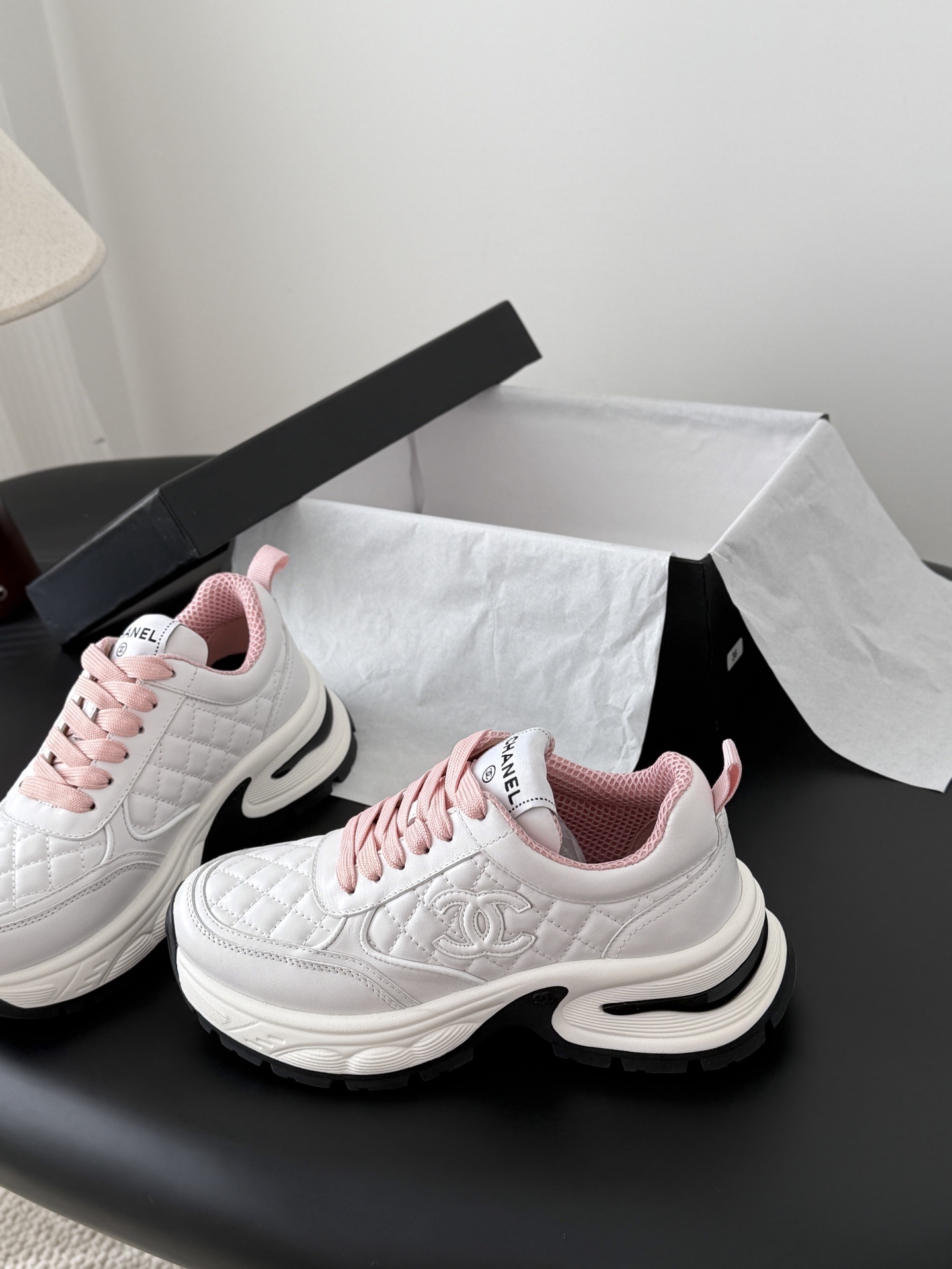 A pair of luxury white Chanel sneakers with pink laces and quilted leather detailing displayed next to a black Chanel shoe box. - Hình 2