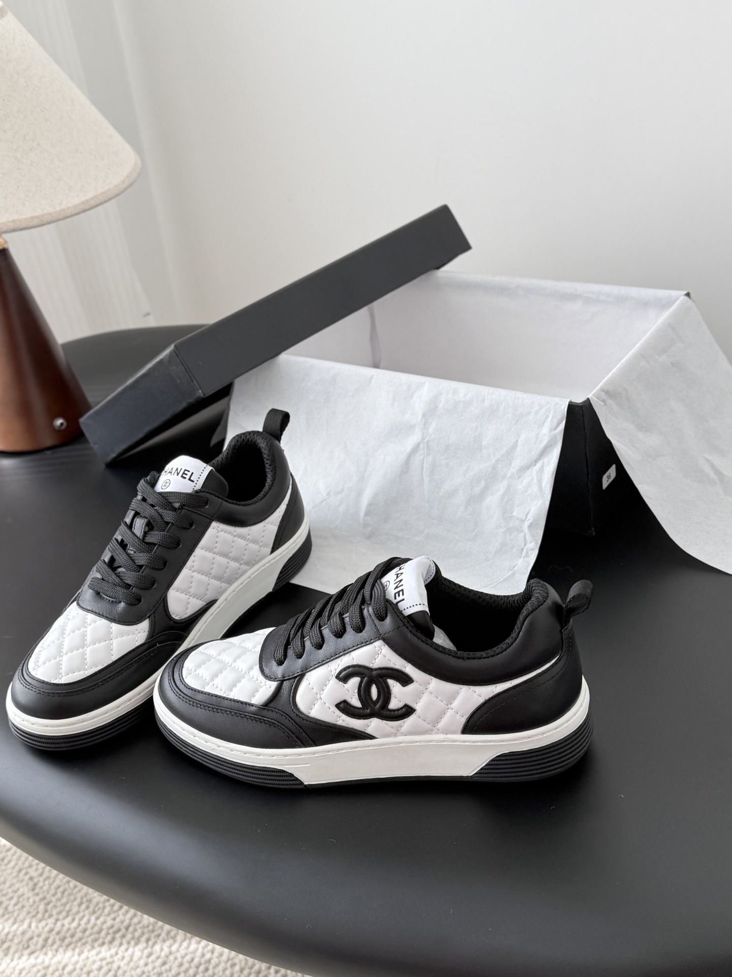 A pair of luxury Chanel black and white quilted leather sneakers with CC logo next to their original black box. - Hình 2