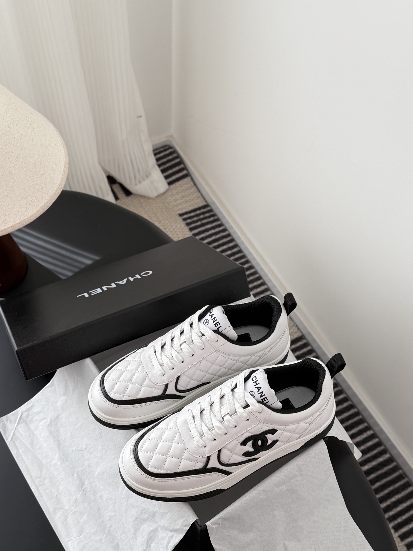 A pair of luxury Chanel white quilted leather sneakers with black CC logo and black designer shoebox on a table. - Hình 9