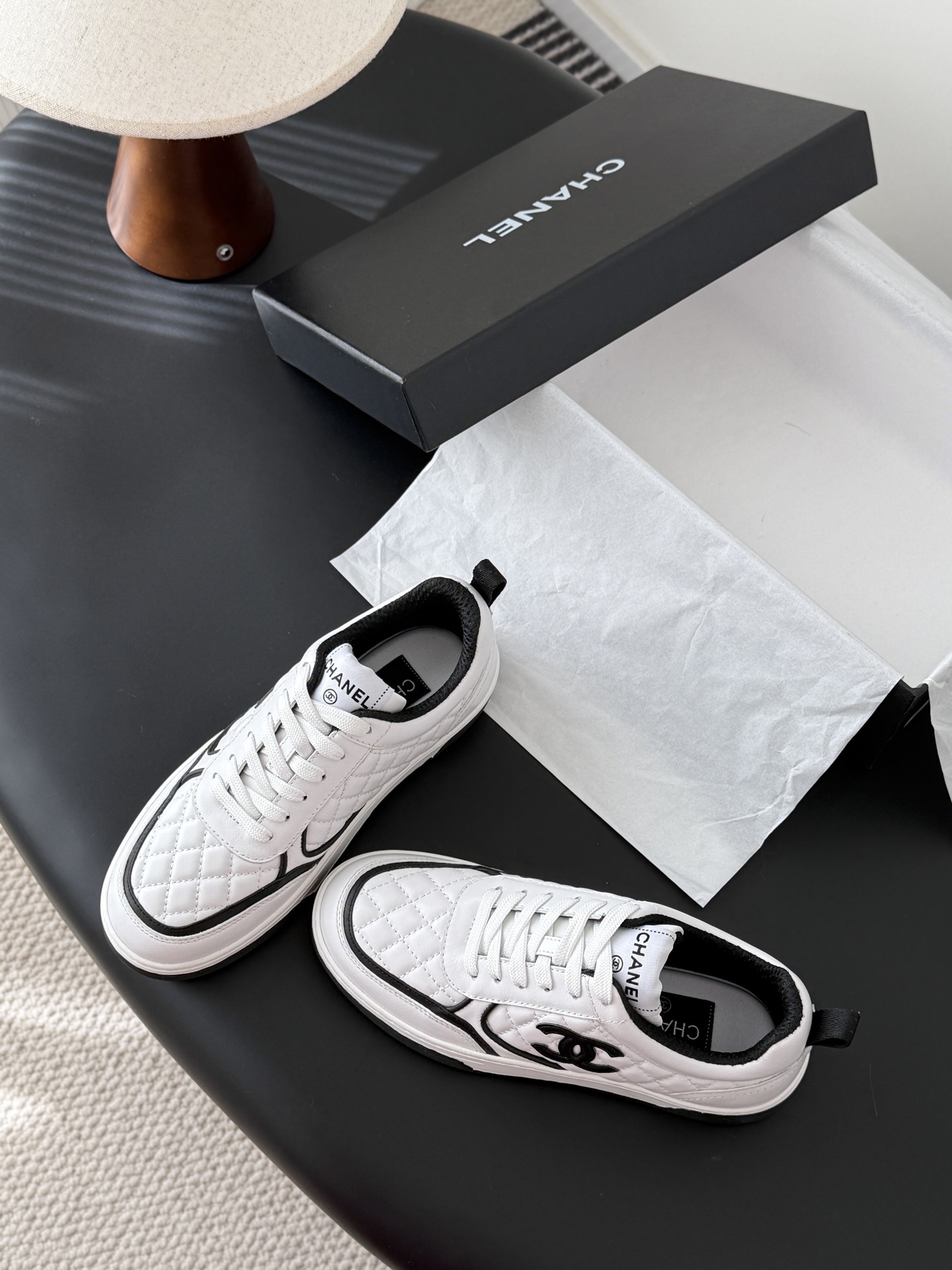 A pair of luxury Chanel white quilted leather sneakers with black CC logo and black designer shoebox on a table. - Hình 6