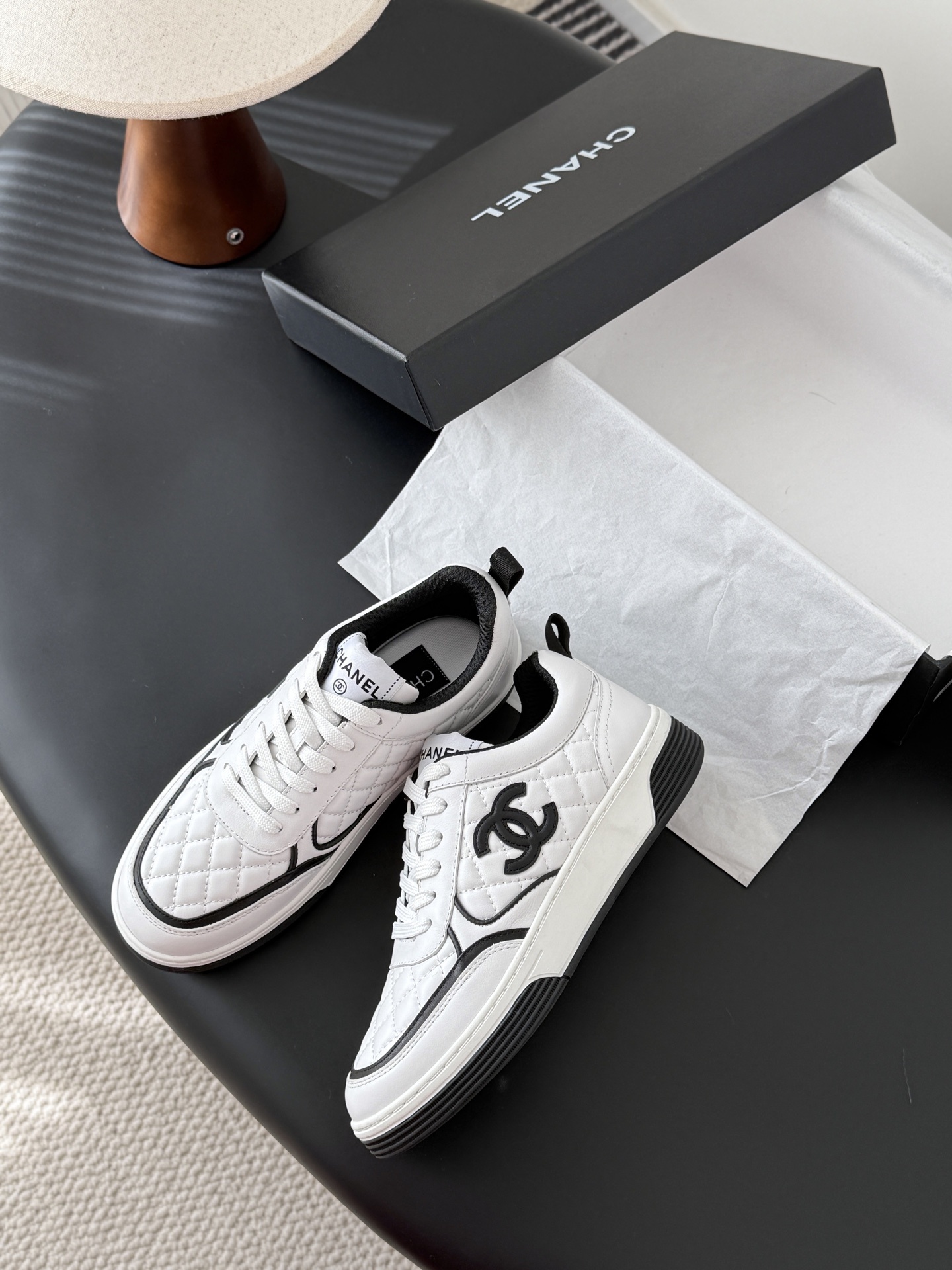 A pair of luxury Chanel white quilted leather sneakers with black CC logo and black designer shoebox on a table. - Hình 4