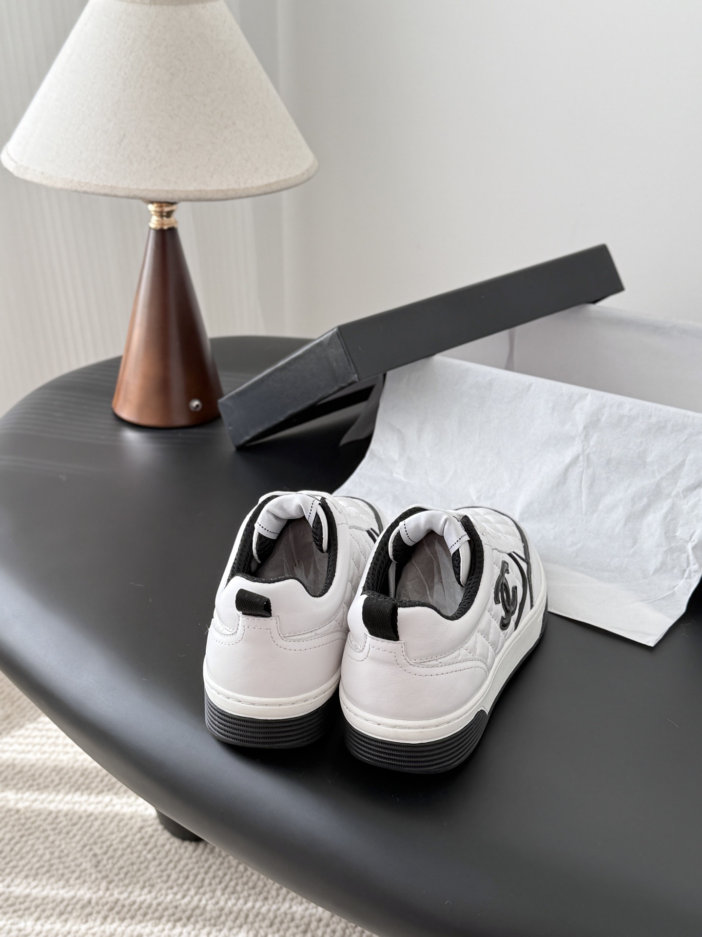 A pair of luxury Chanel white quilted leather sneakers with black CC logo and black designer shoebox on a table. - Hình 3