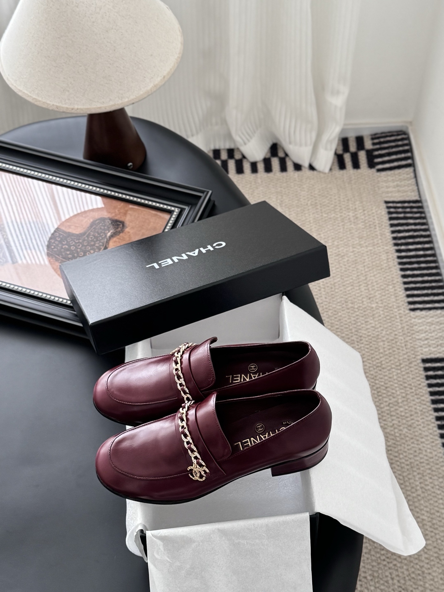 Pair of burgundy Chanel leather loafers with gold chain detail and block heels next to a black Chanel shoe box. - Hình 9